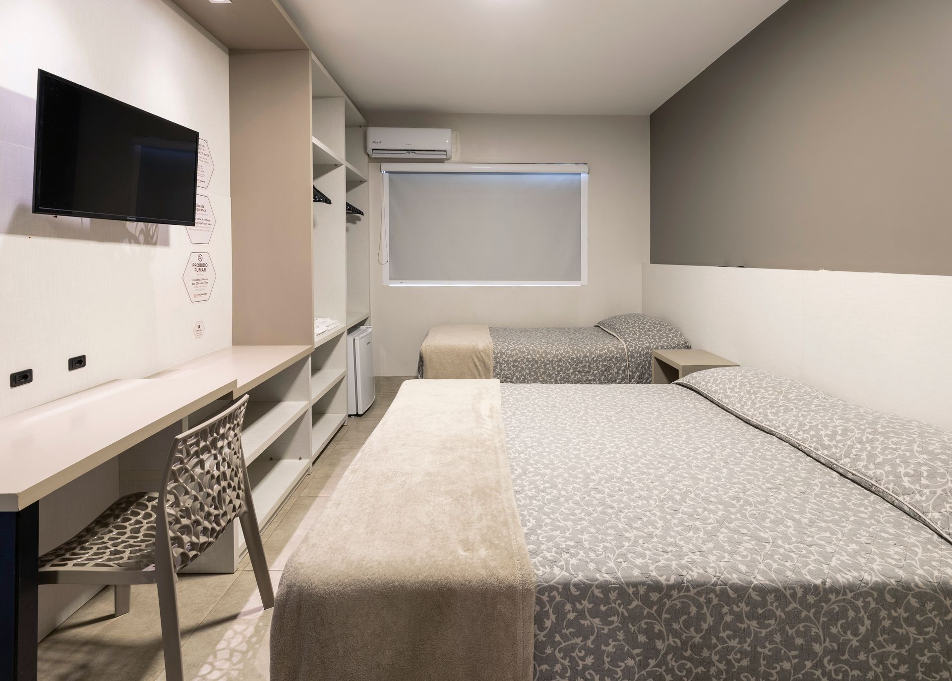 Quarto de hotel moderno com duas camas, TV de parede, escrivaninha, cadeira e persianas.