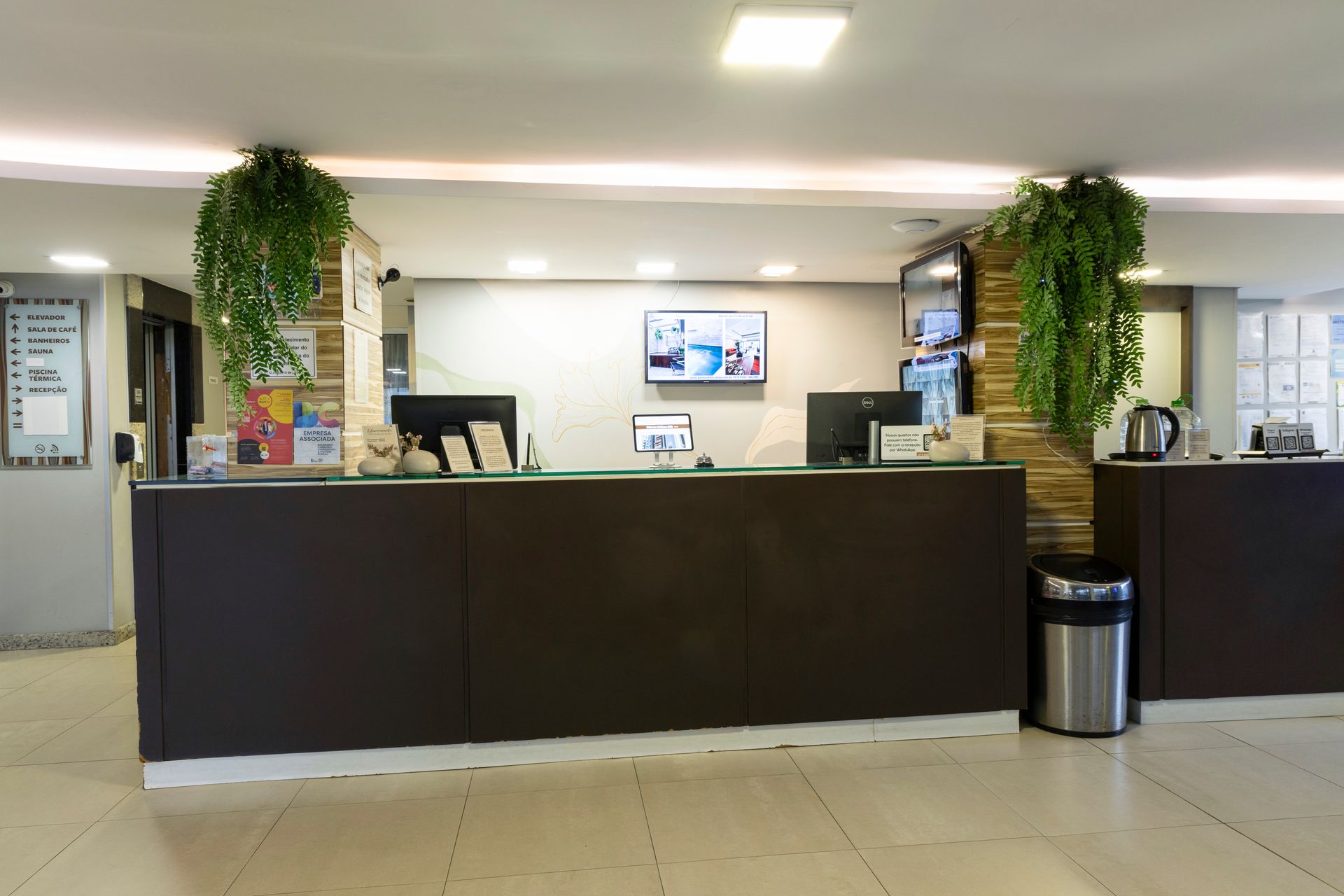 Balcão de recepção do hotel com balcões escuros, plantas suspensas e uma TV ao fundo.
