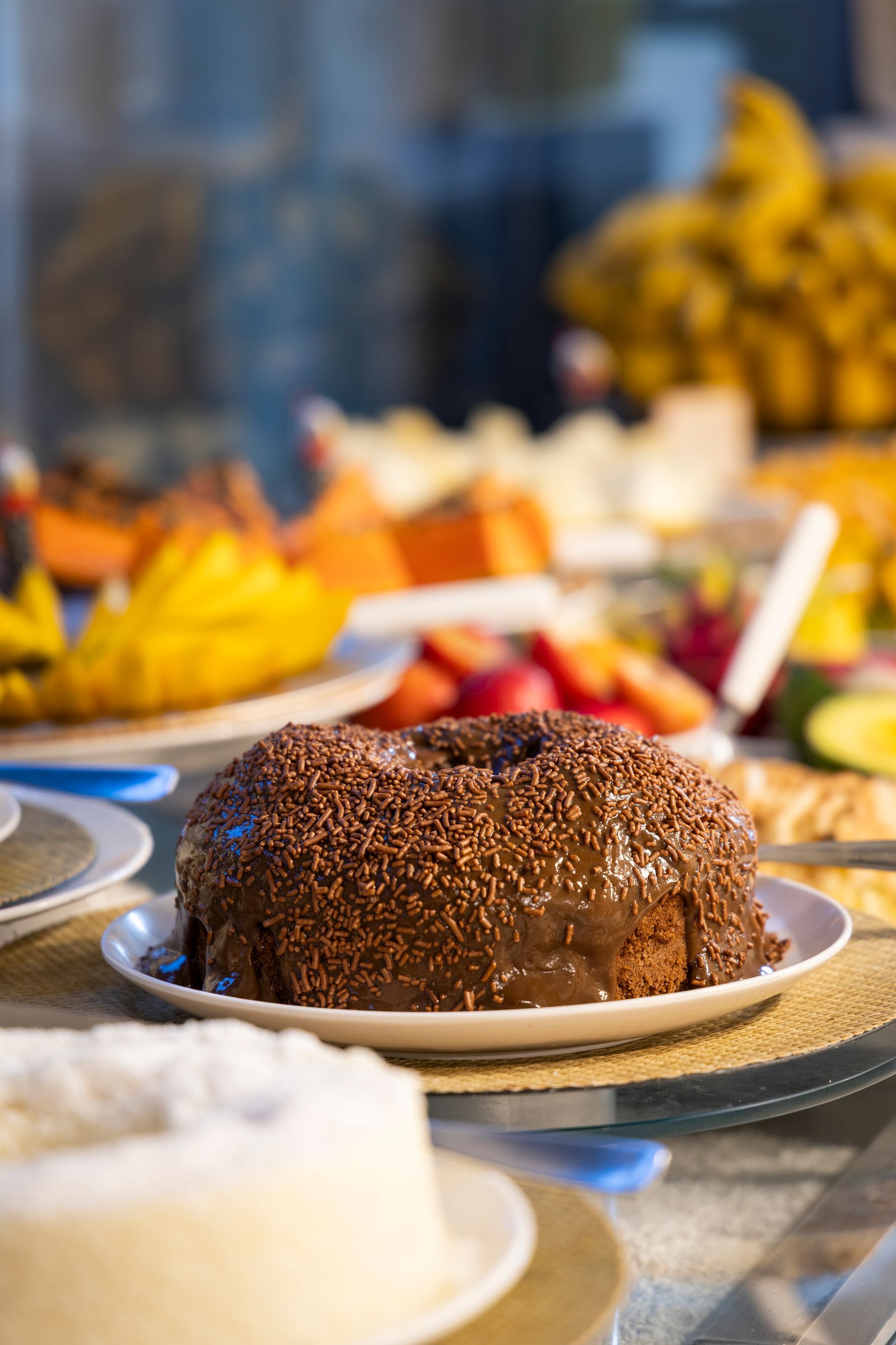 Bolo de chocolate em um prato em uma mesa de banquete com frutas e sobremesas ao fundo.
