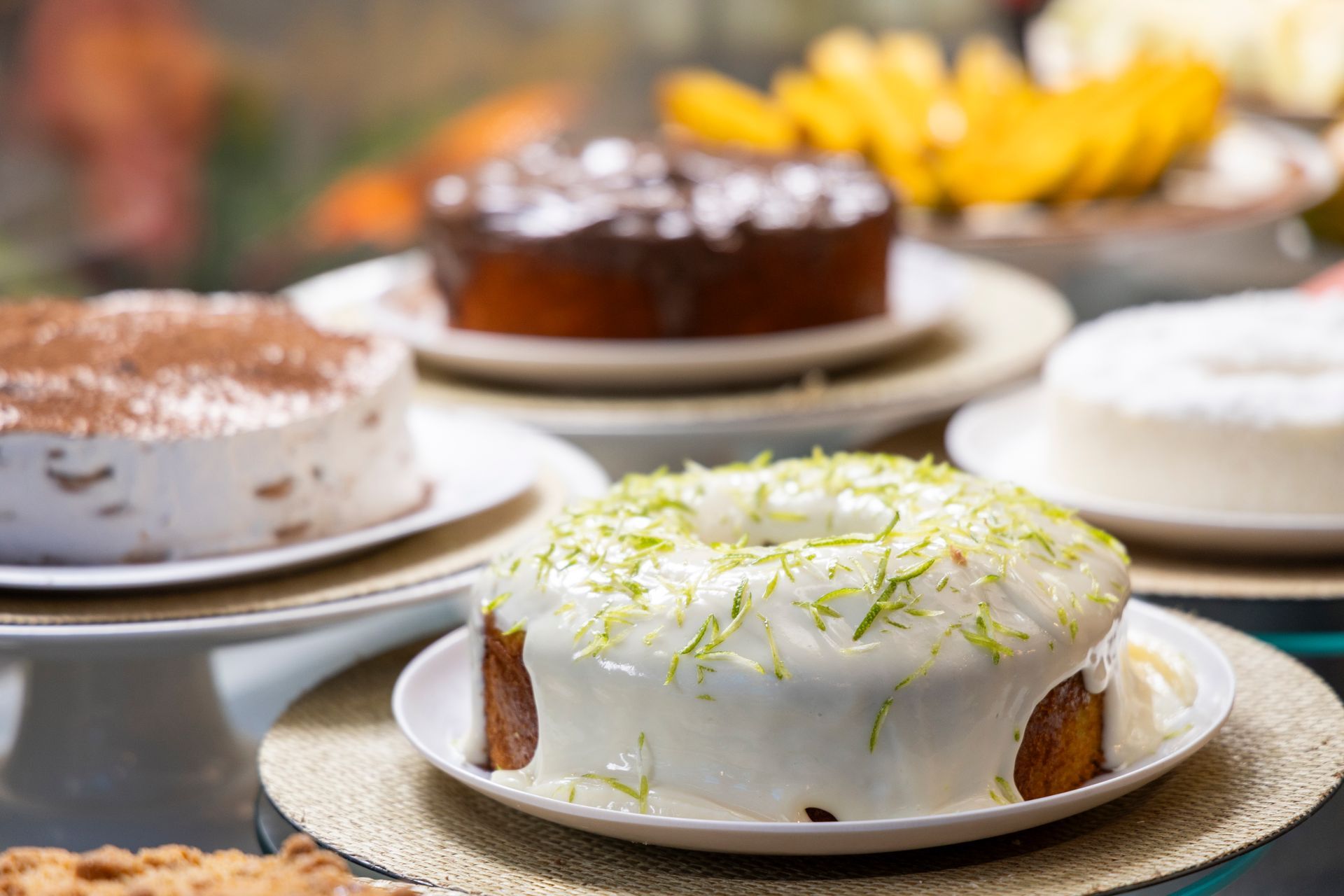 Diversos bolos confeitados dispostos em pratos, com um bolo de cobertura branca e raspas de limão em primeiro plano.