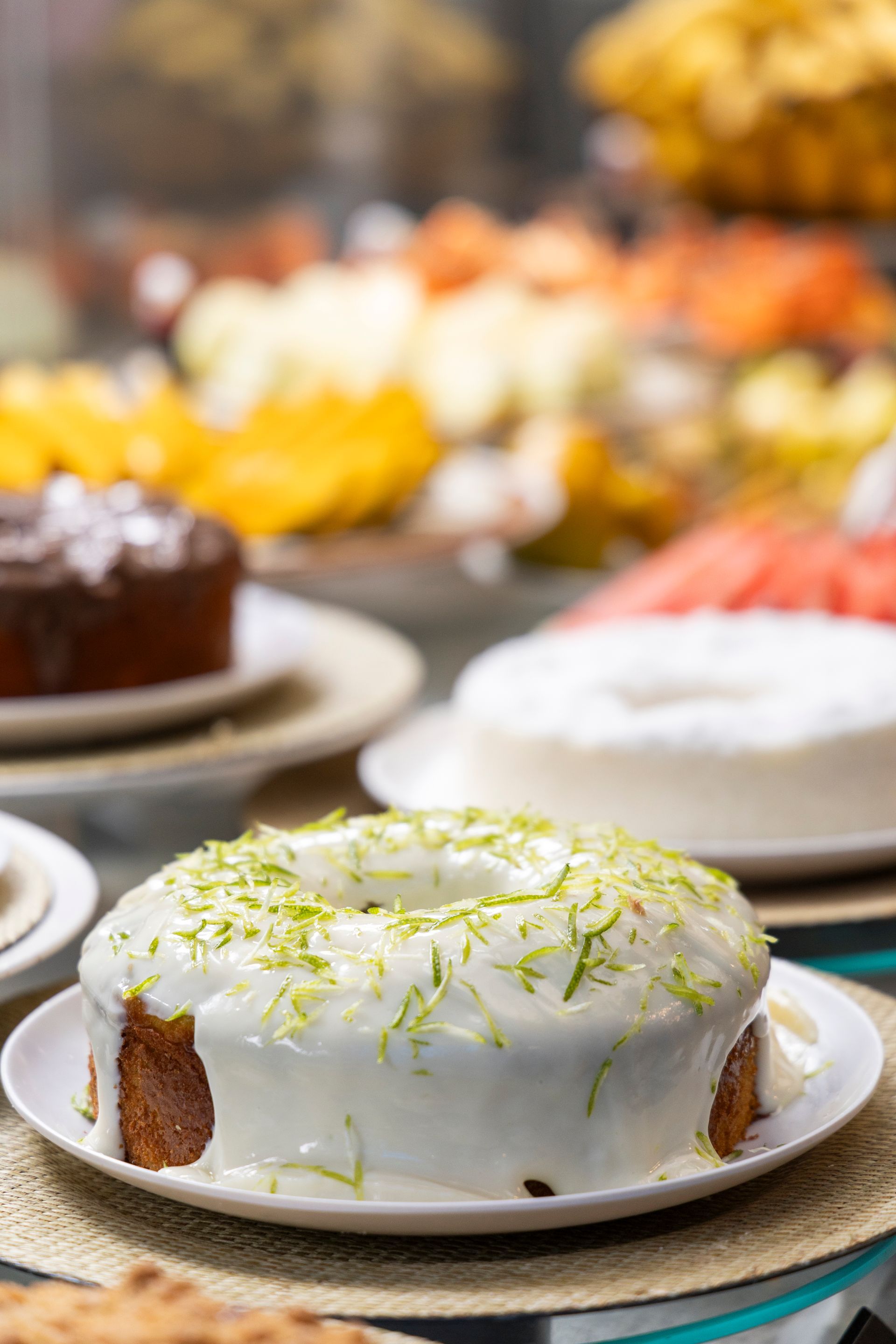 Bolo Bundt com cobertura de limão e raspas de limão em um prato, com outros bolos desfocados ao fundo.