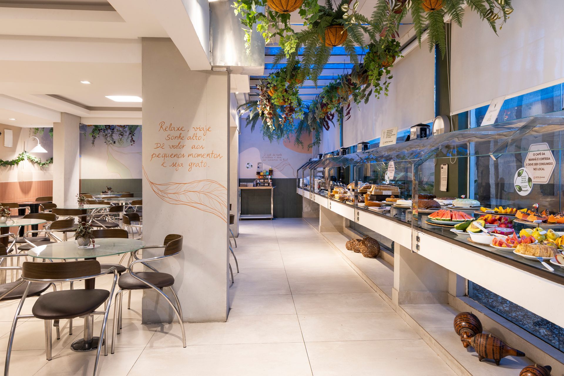 Cafeteria moderna com mesas, balcão de buffet e plantas suspensas sob luzes brilhantes.