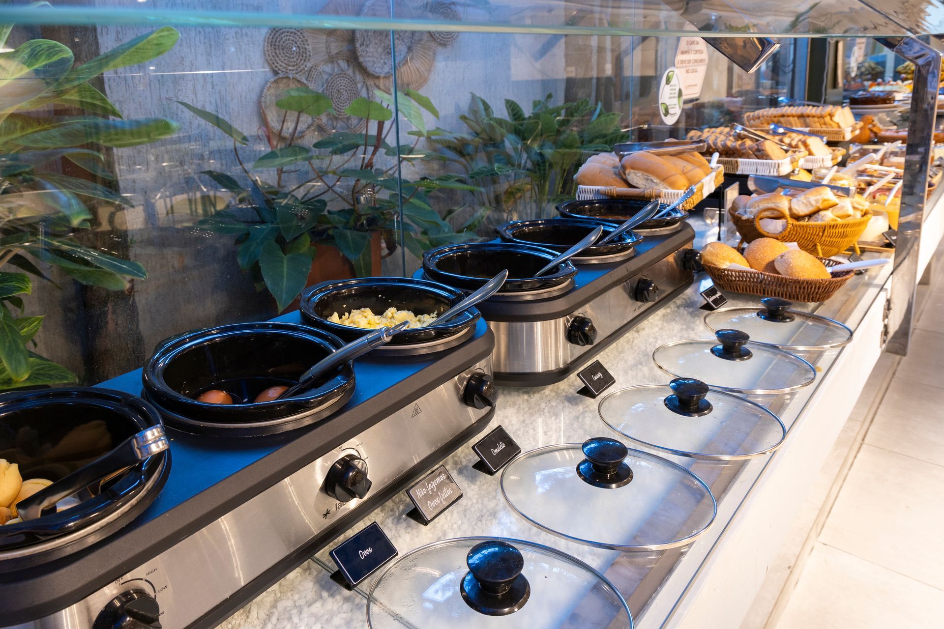 Buffet self-service com pratos cobertos e uma fileira de doces em um balcão de aço inoxidável.