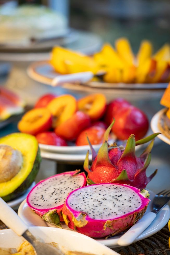 Uma variedade de frutas tropicais dispostas sobre uma mesa, incluindo fatias de pitaya, manga, abacate, morangos e maçãs vermelhas.