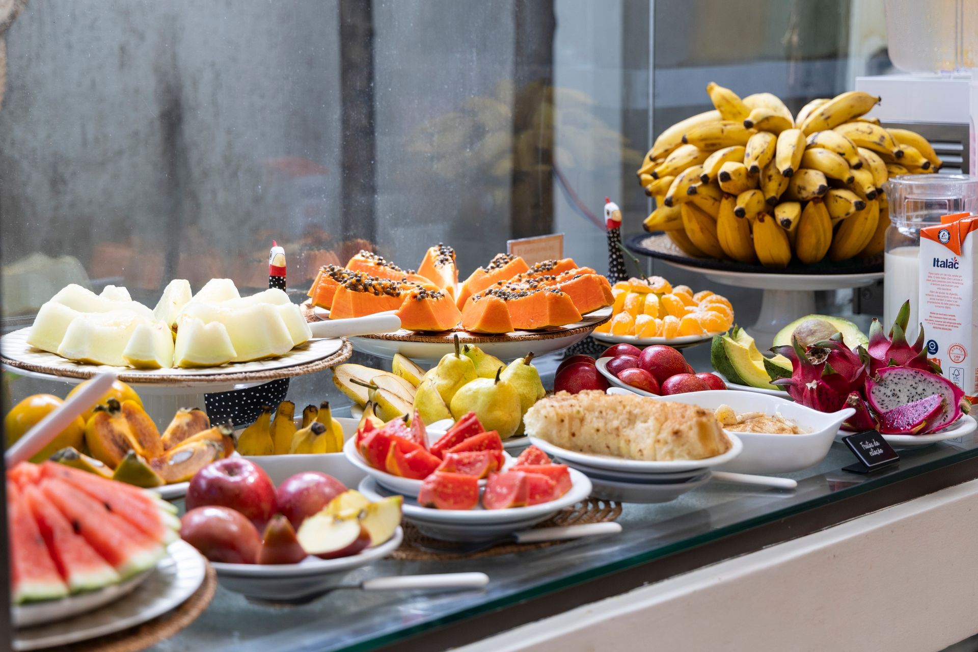 Mesa de buffet com travessas variadas de frutas, queijos, doces e pães, dispostas em um ambiente de restaurante.