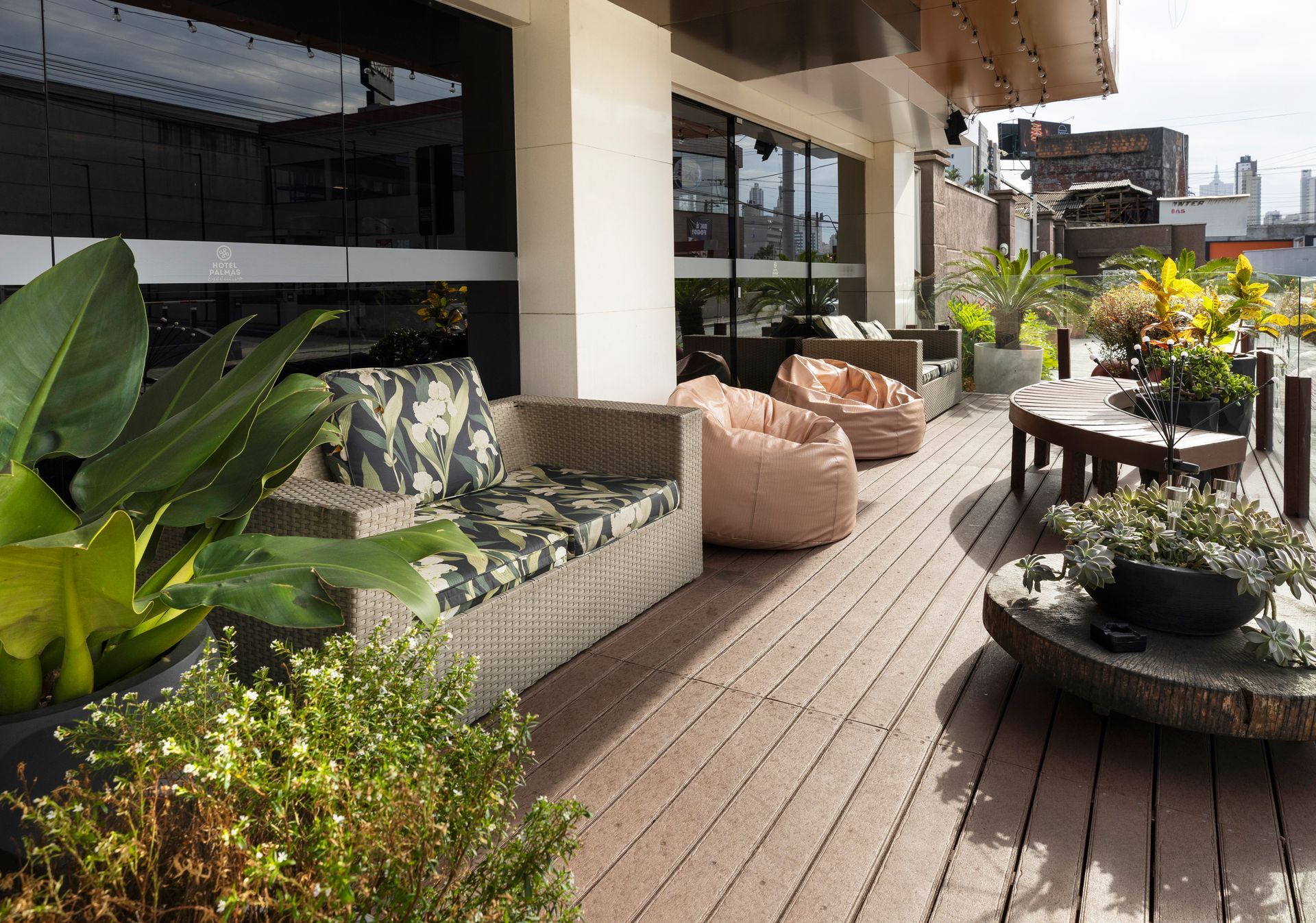 Pátio externo ensolarado com deck de madeira, assentos acolchoados, plantas em vasos e vista para os edifícios próximos.