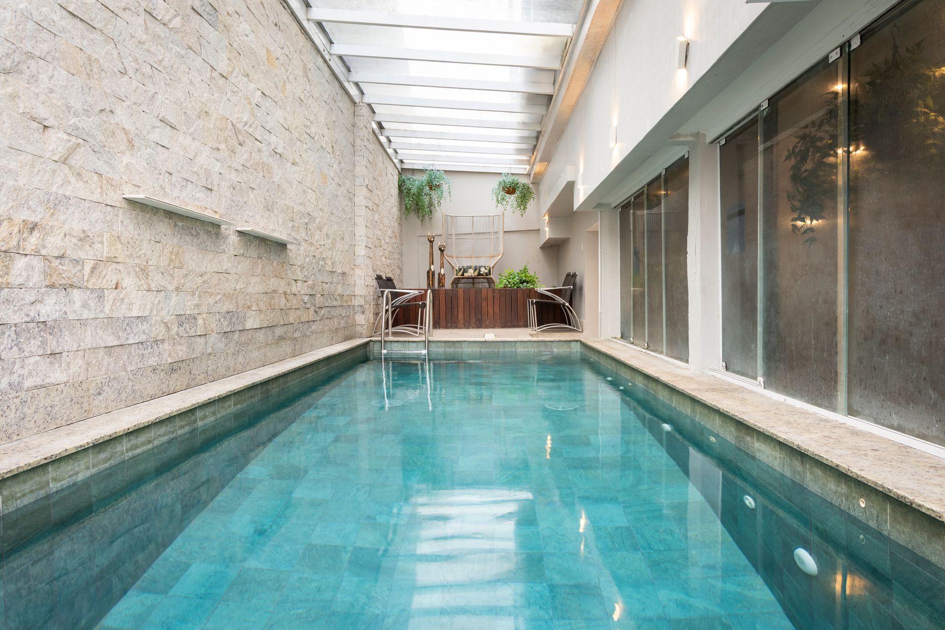 Piscina coberta para natação com água cristalina turquesa ao lado de paredes de pedra e vidro sob uma claraboia.