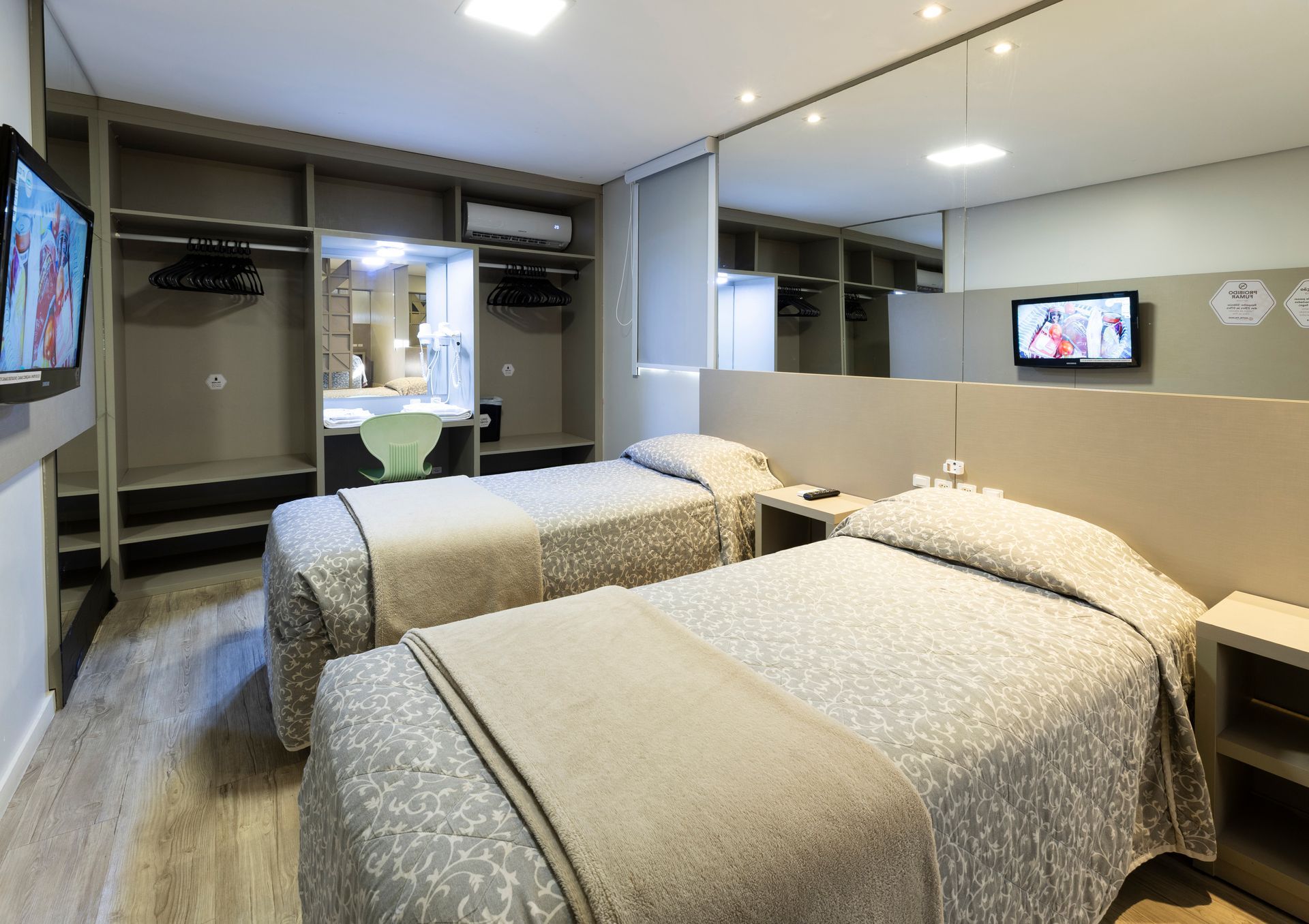 Quarto de hotel moderno com duas camas de solteiro, armário com espelho e TV de parede em um ambiente iluminado.