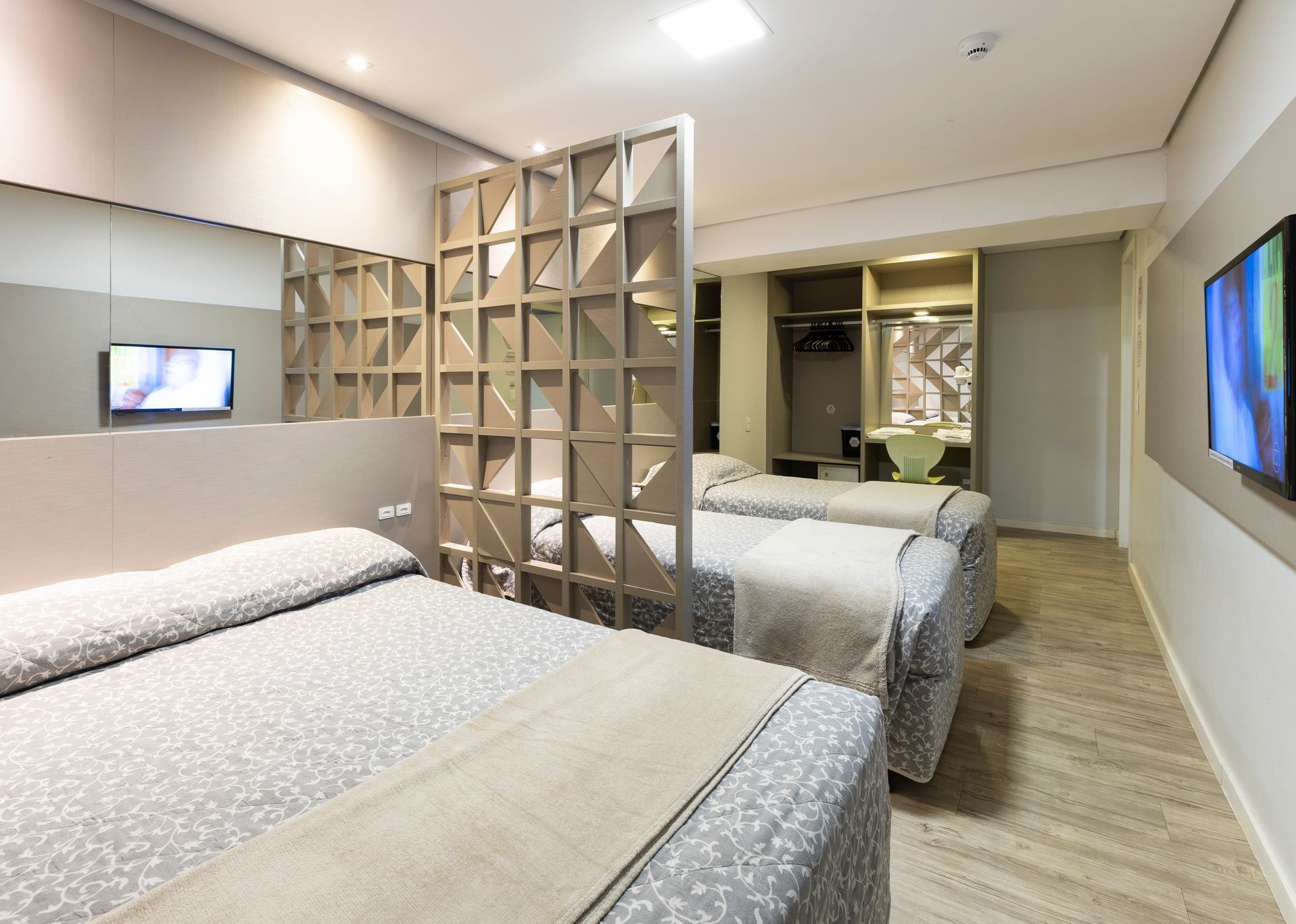 Quarto de hotel pequeno com duas camas, um guarda-roupa com espelho, televisões fixadas na parede e piso de madeira.