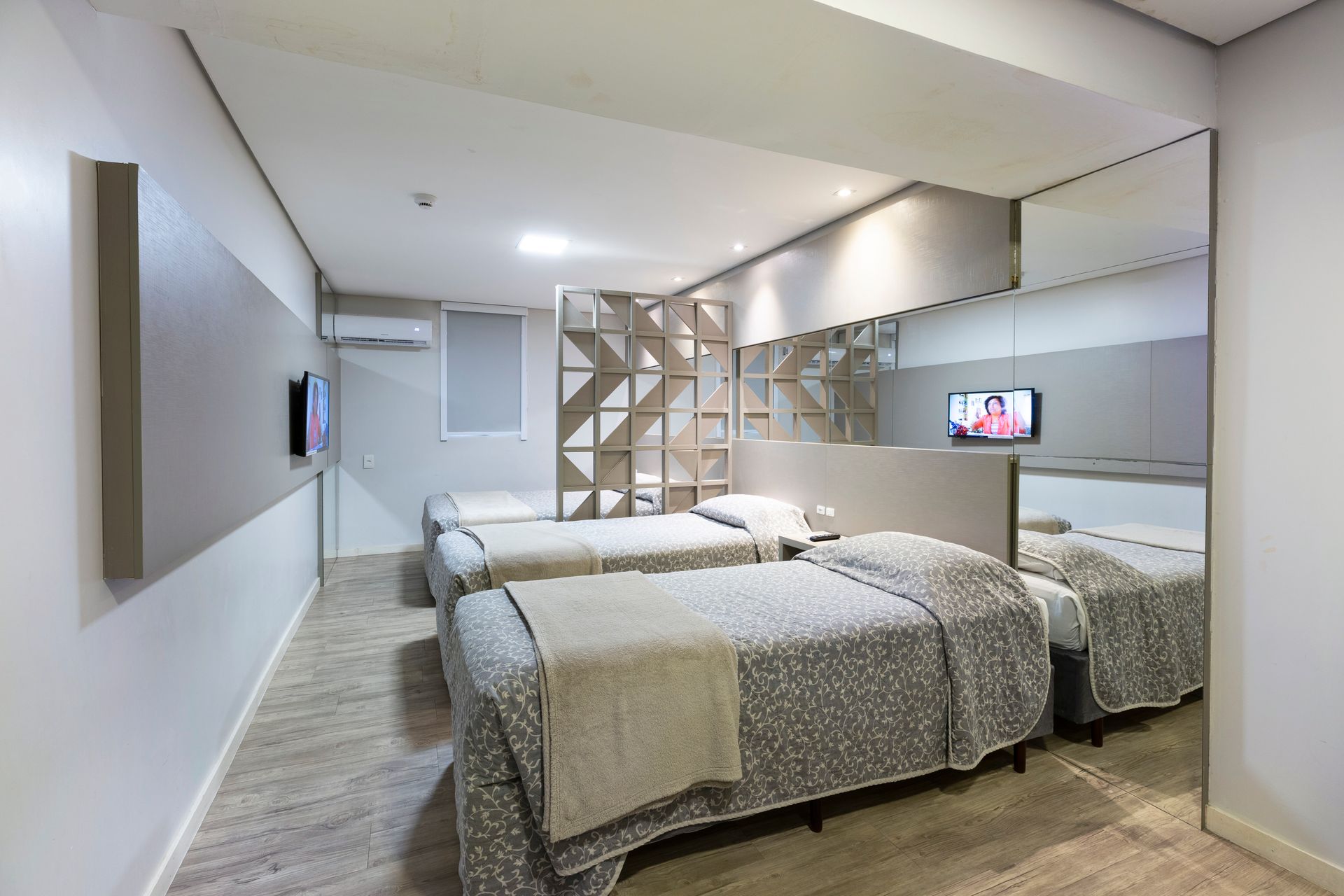 Quarto de hotel moderno com duas camas, espelho de parede, cabeceira geométrica e TV de parede.