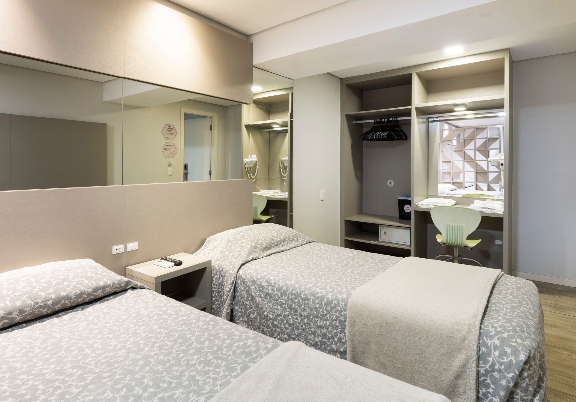 Quarto de hotel pequeno com duas camas de solteiro, parede espelhada e uma escrivaninha junto à janela.