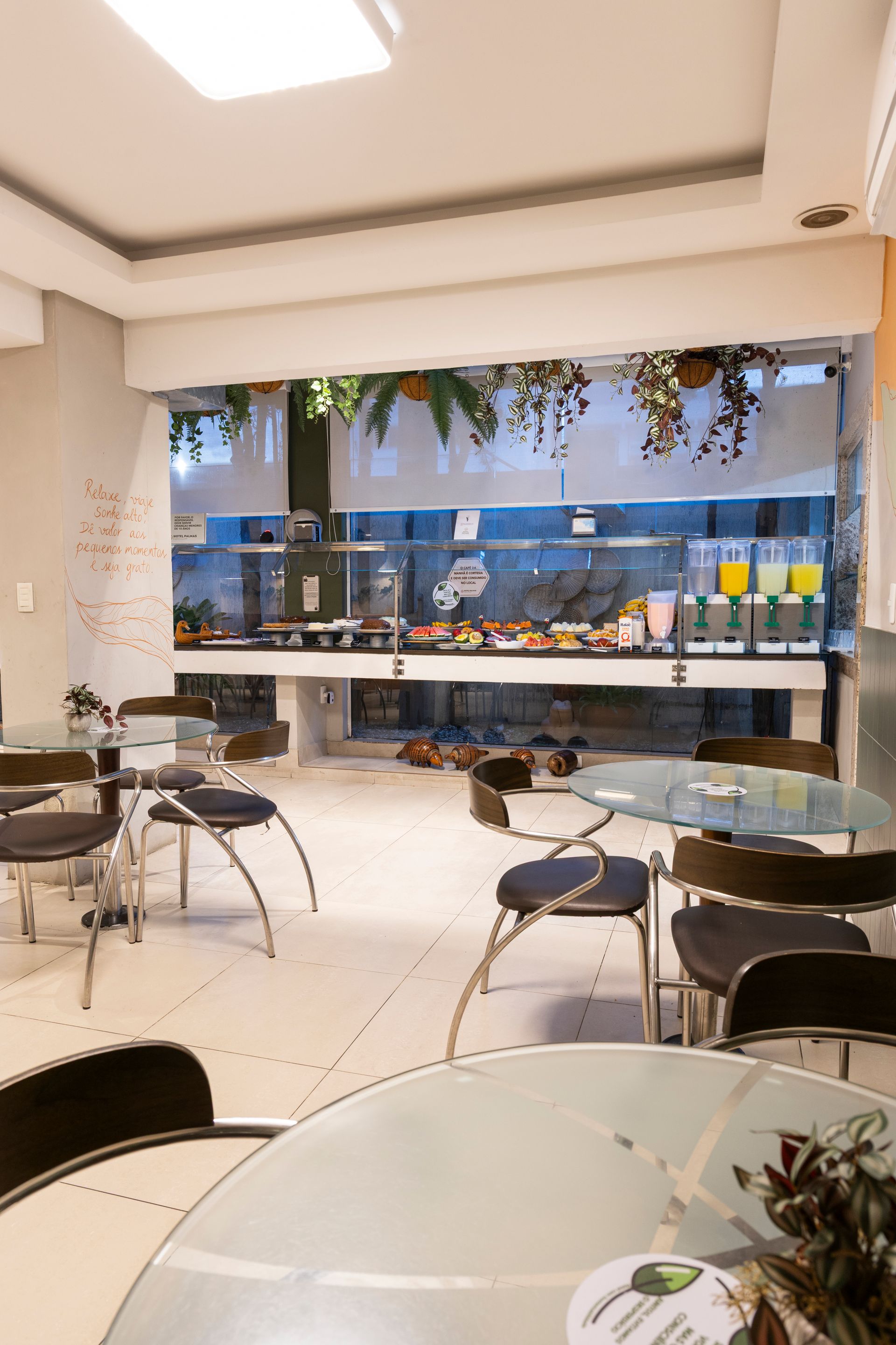 Interior de café moderno com mesas redondas, cadeiras pretas e um longo balcão decorado com folhagens suspensas.