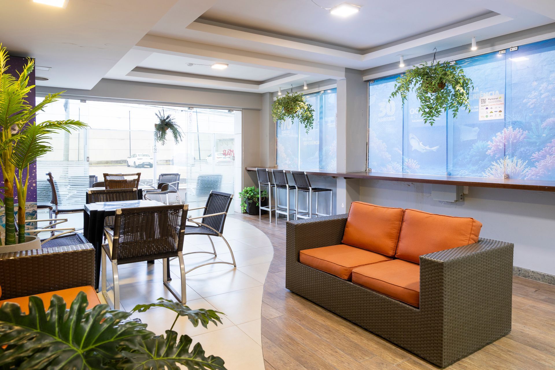 Sala de estar moderna com sofá laranja, cadeiras de vime e plantas suspensas em um saguão de escritório iluminado.