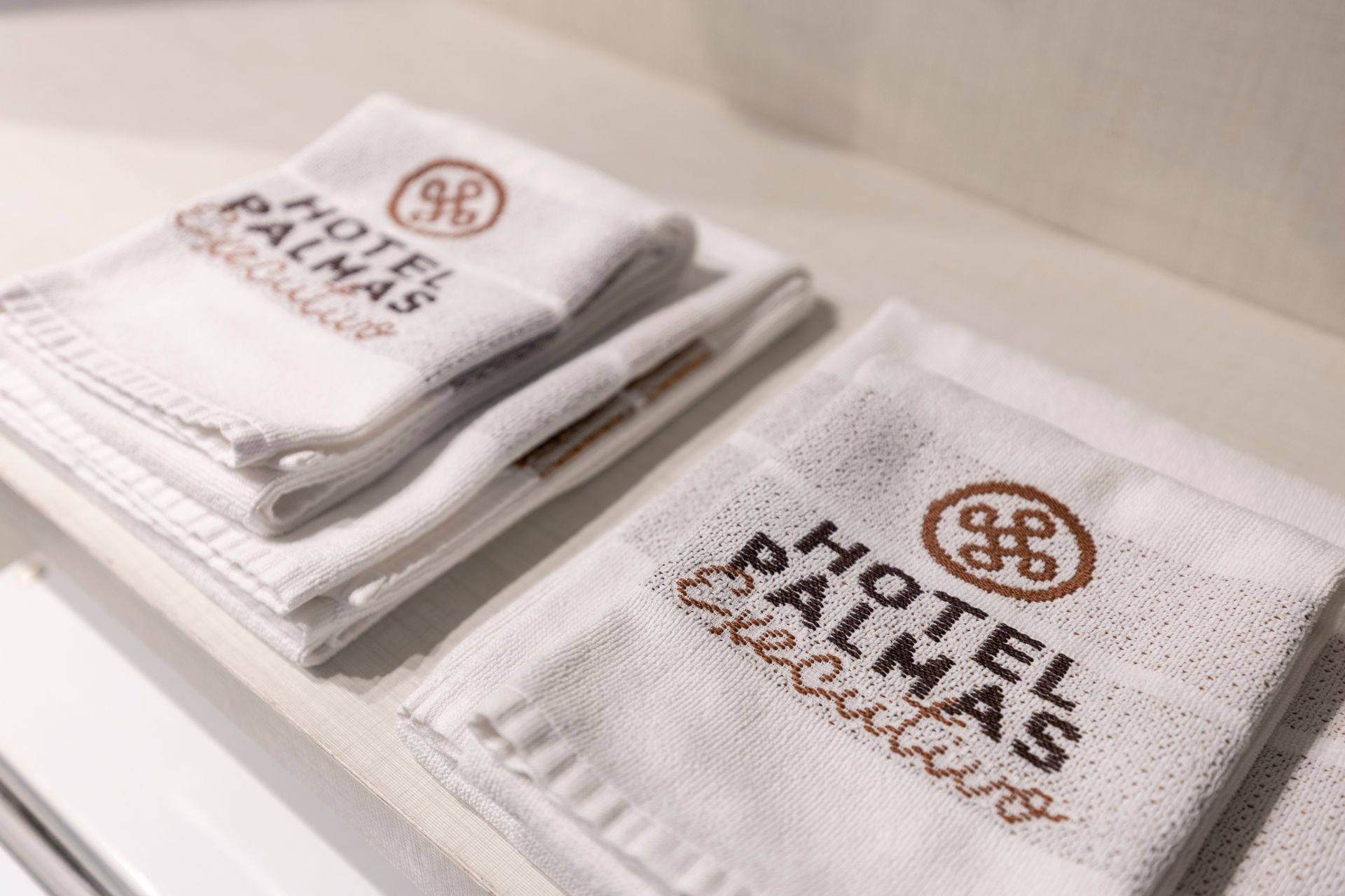 Toalhas brancas dobradas e empilhadas, com logotipos bordados, sobre uma prateleira.