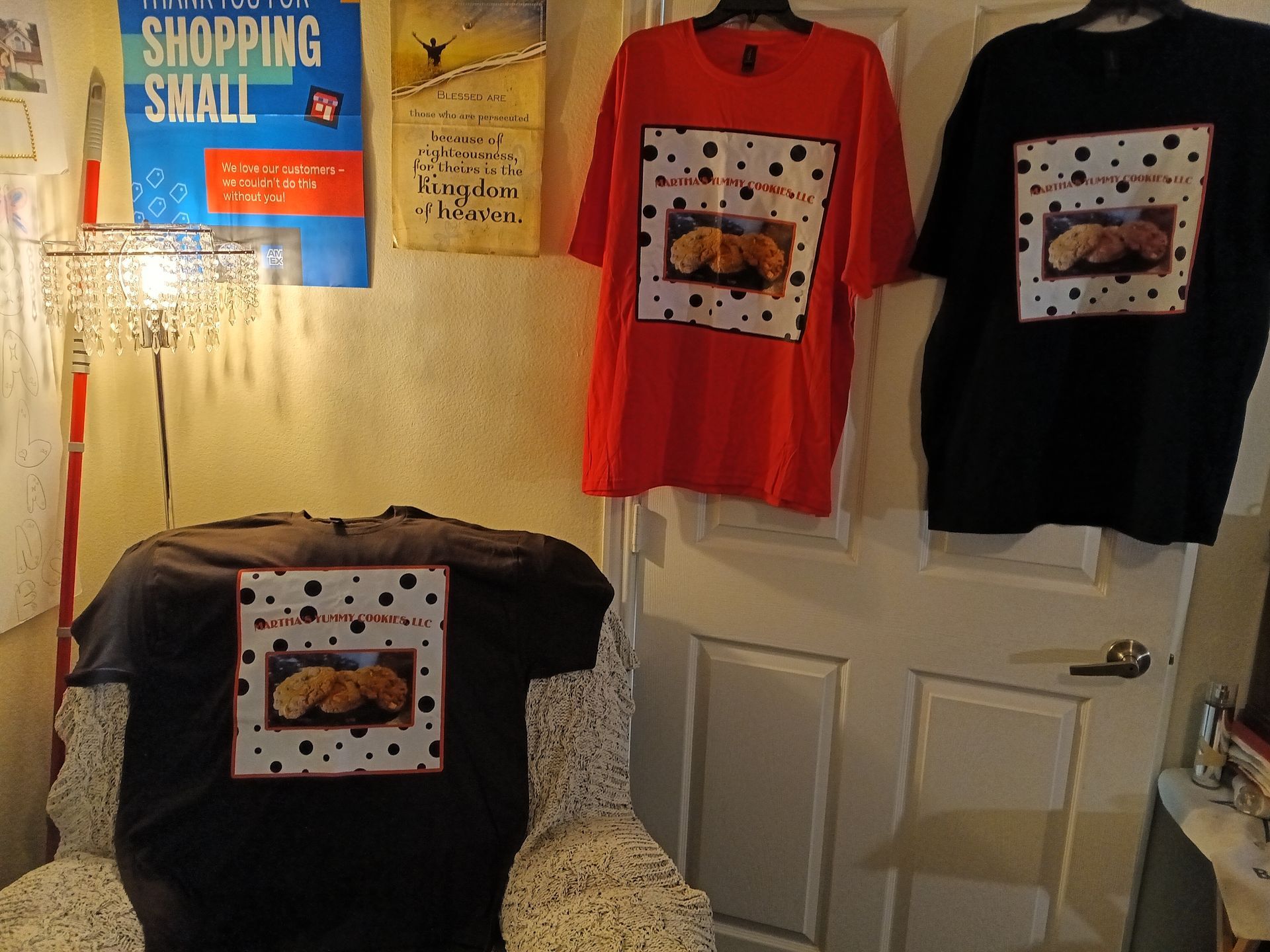 Three identical t-shirts featuring a graphic of cookies, displayed on a chair and hung on a door, with a poster nearby.