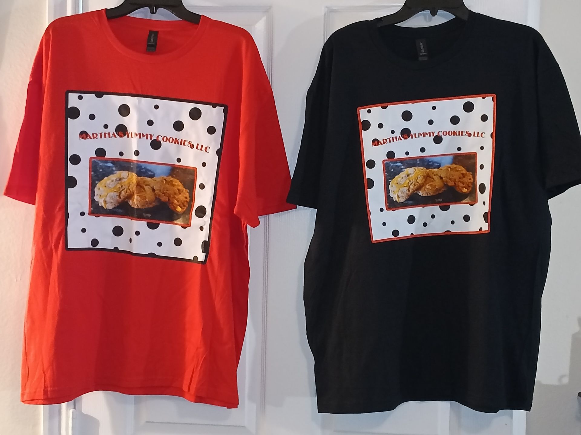 A red shirt and a black shirt, both featuring a square print of chocolate chip cookies on a polka-dot background.