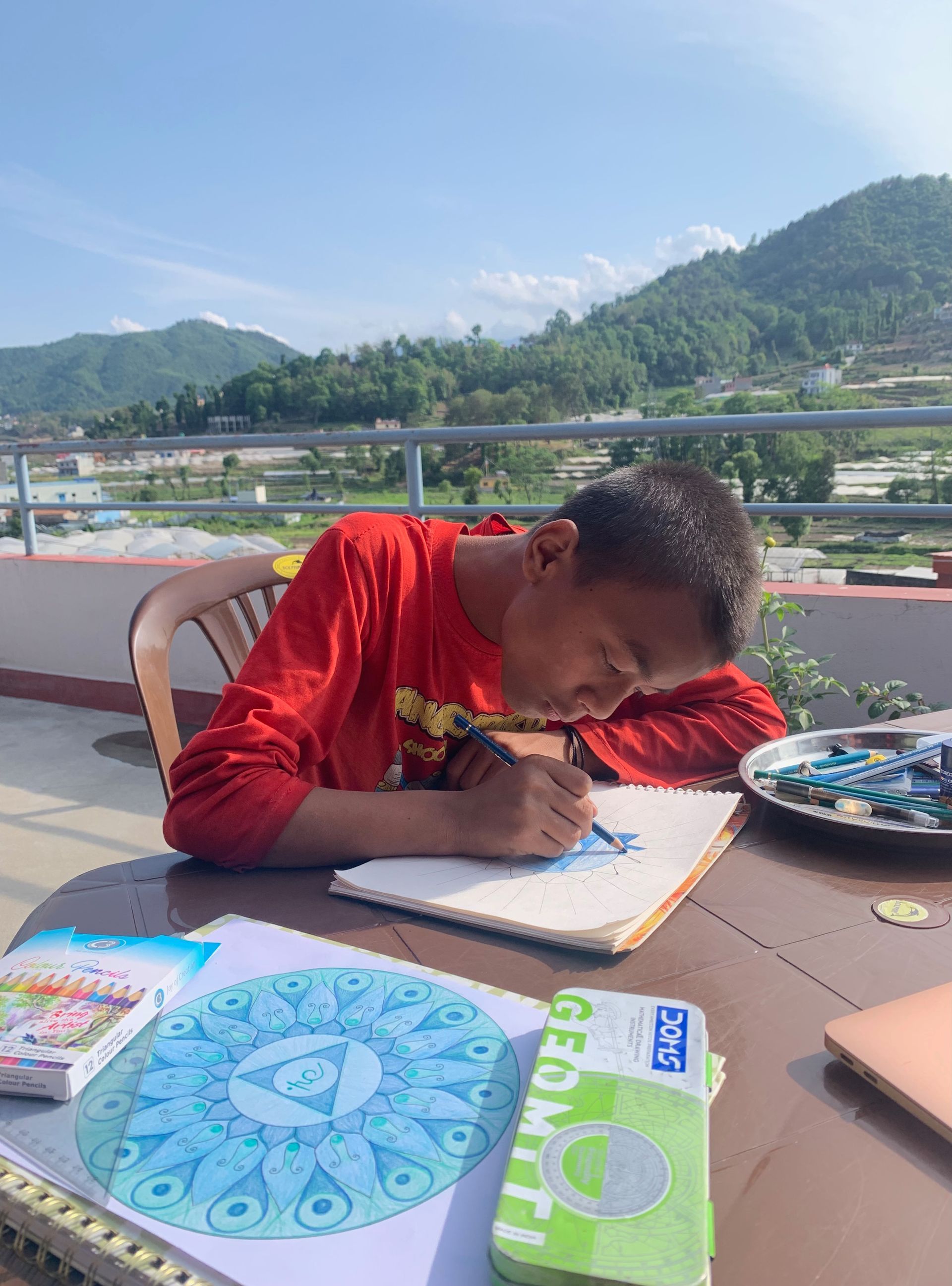 Young person drawing on a rooftop, mountains in the background. Wearing red shirt, using blue/green colors, sunny day.