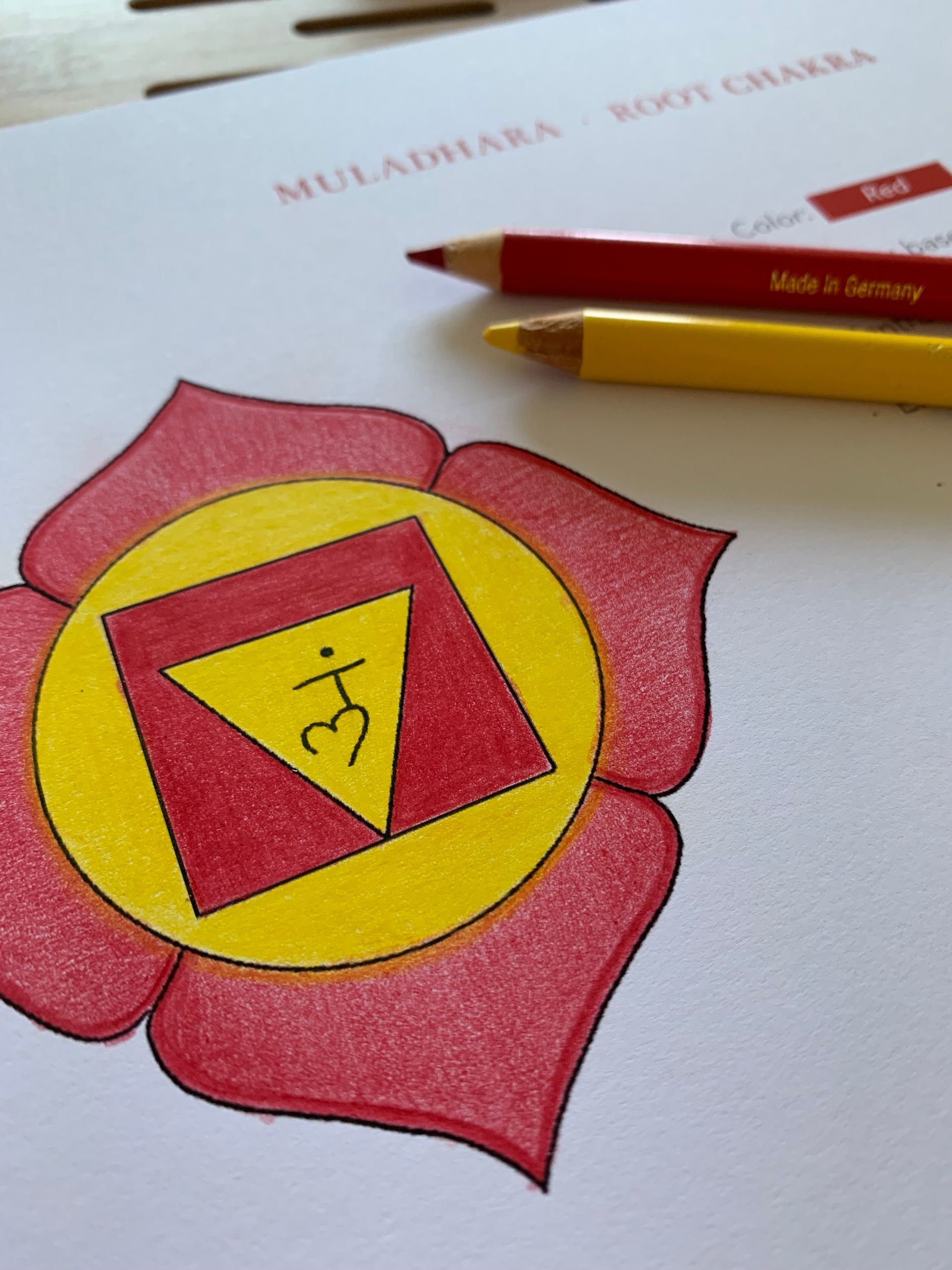 A red and yellow chakra diagram being colored with red and yellow pencils; text indicates it's the root chakra.