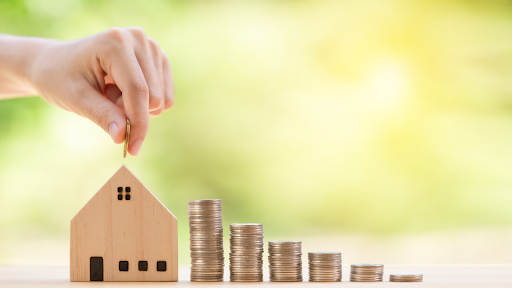 A hand is placing a coin into a small wooden house-shaped model, symbolizing saving or investing in real estate.