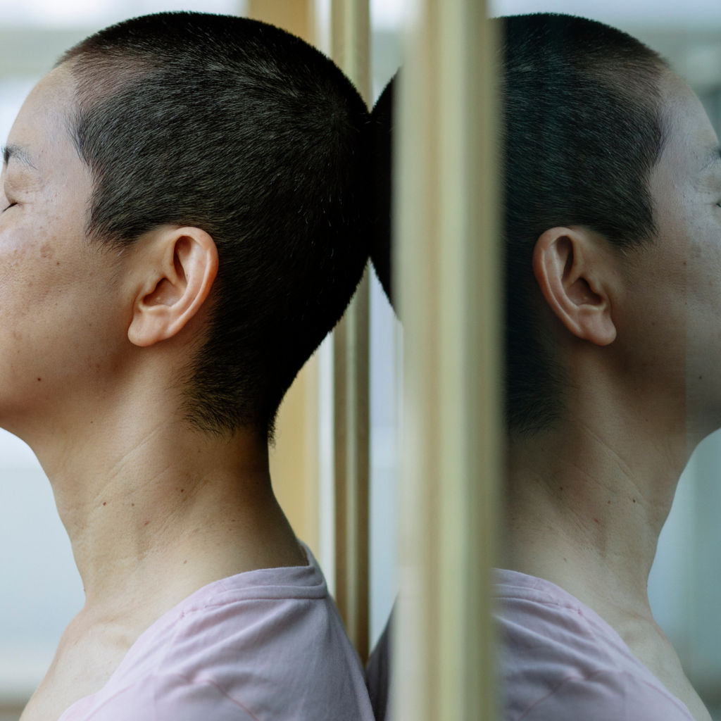 A woman with a shaved head is looking at her reflection in a mirror