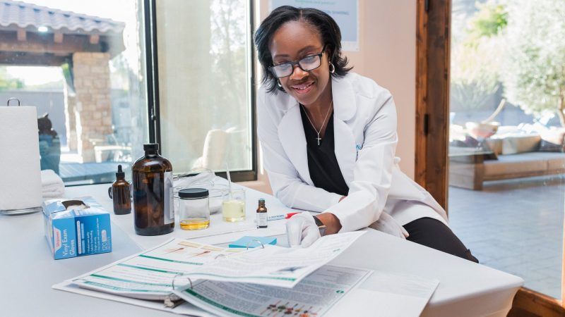 A woman in a lab coat is sitting at a table with bottles of medication.