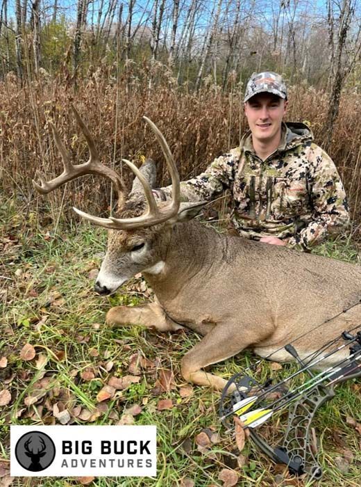 A man is kneeling next to a deer with a bow in the grass.