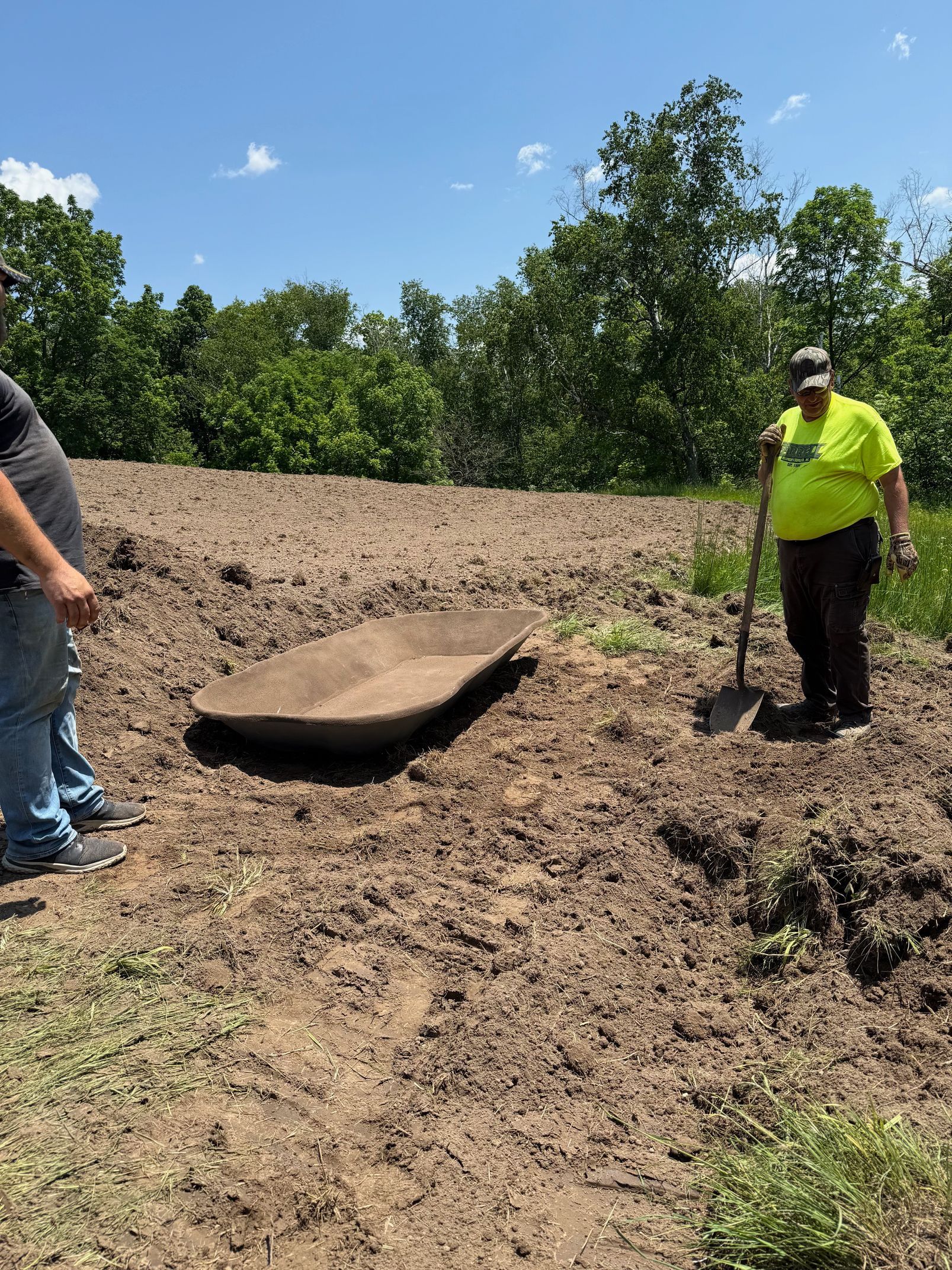Two men are digging in a field with a shovel and a wheelbarrow.