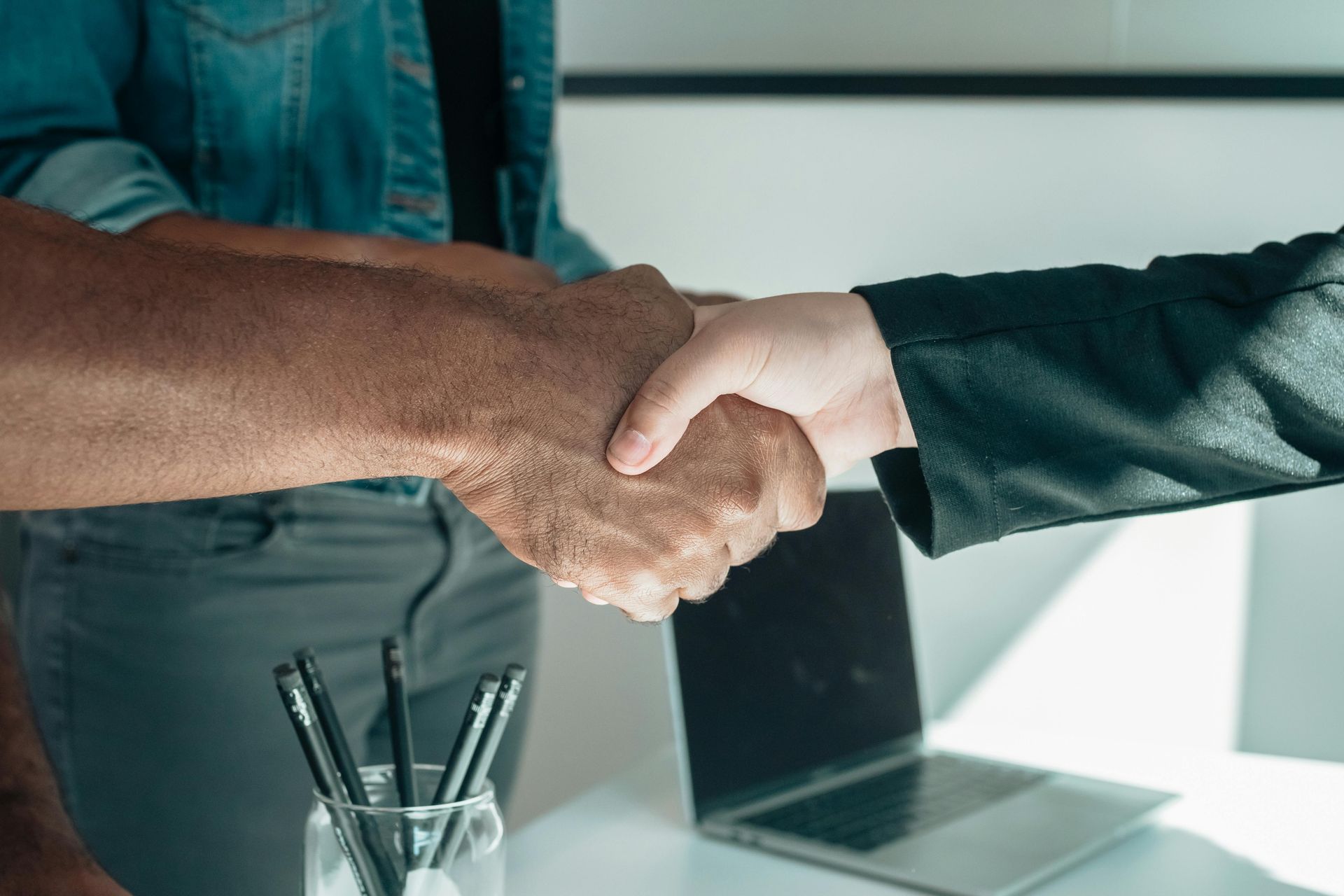 Two people shaking hands over a desk with a laptop and pens, symbolizing agreement or collaboration.