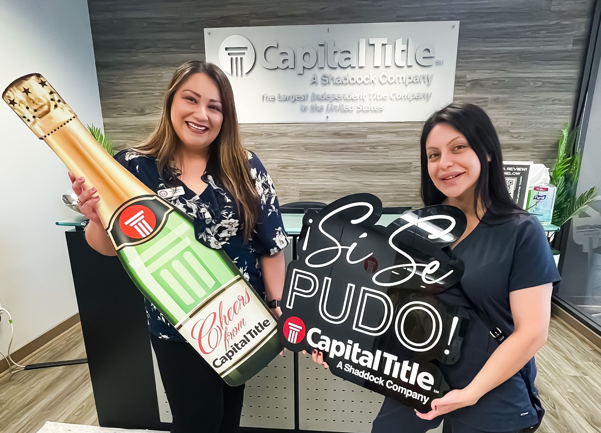 Two women holding celebratory signs in a Capital Title office. One holds a champagne bottle sign, the other says