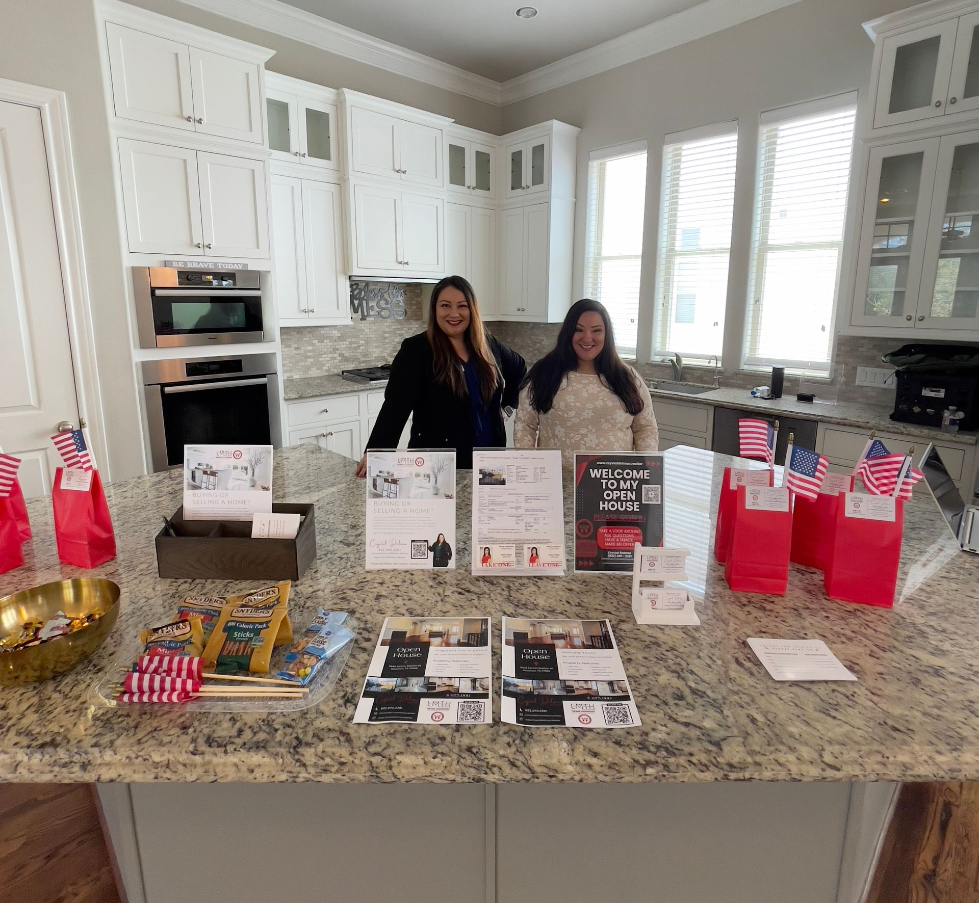 Two women stand behind a kitchen countertop display, showcasing brochures and small gifts. American flags adorn the display.