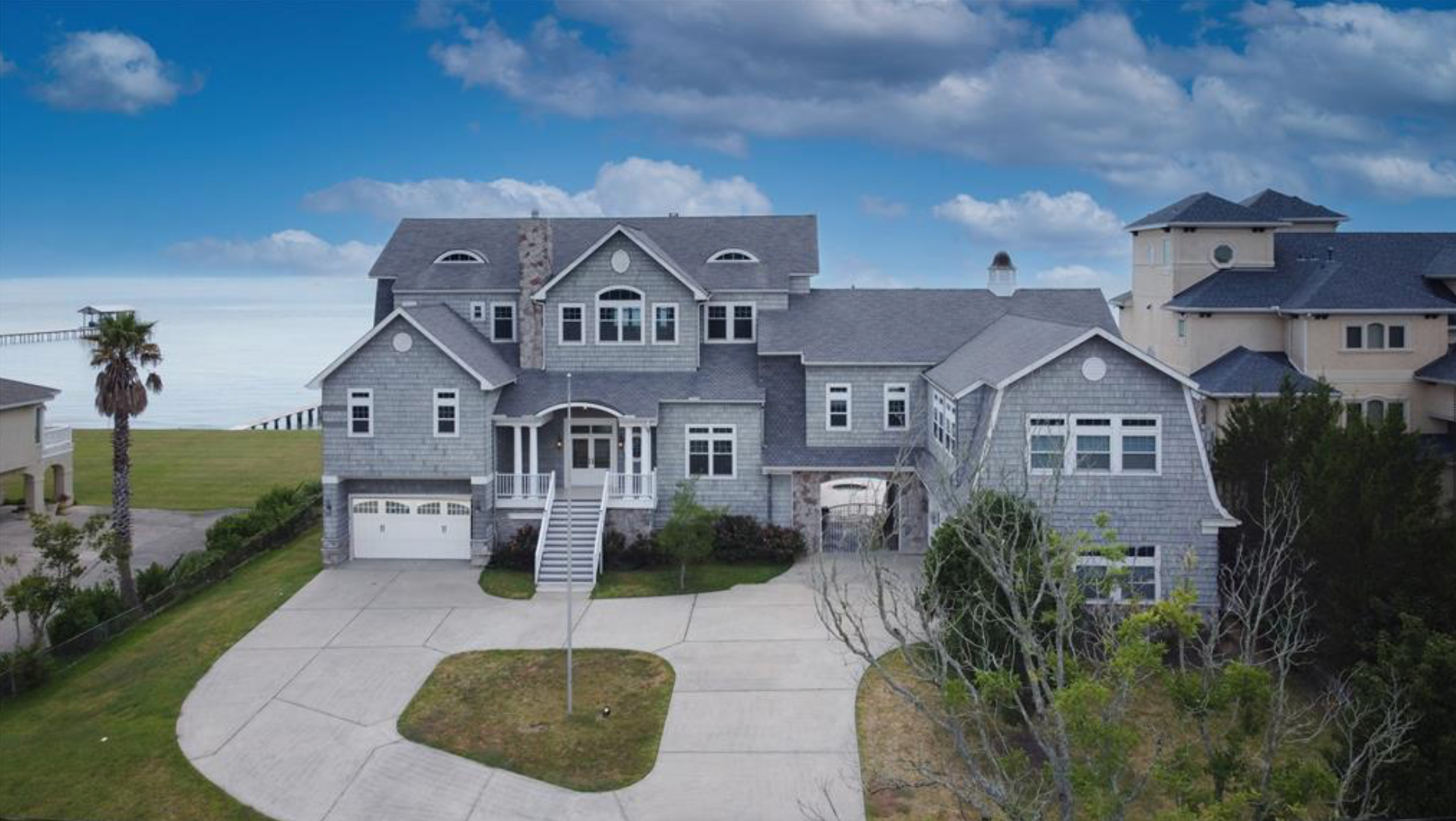 Large gray house with a driveway. Palm tree and ocean in the background. Bright blue sky.