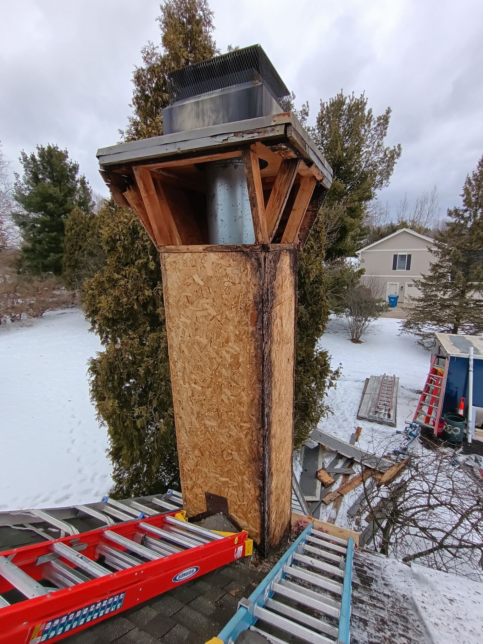 Chimney with a wooden frame and exposed particleboard siding on a snowy roof.