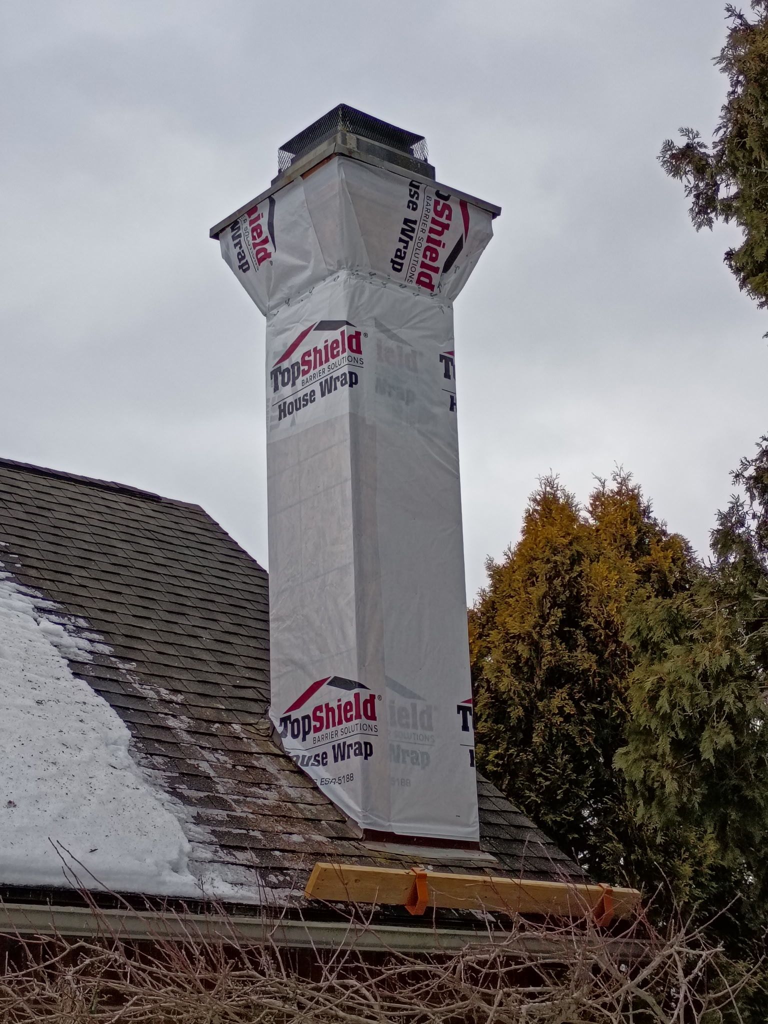 Tall chimney wrapped in protective material, atop a snow-covered roof, cloudy sky.
