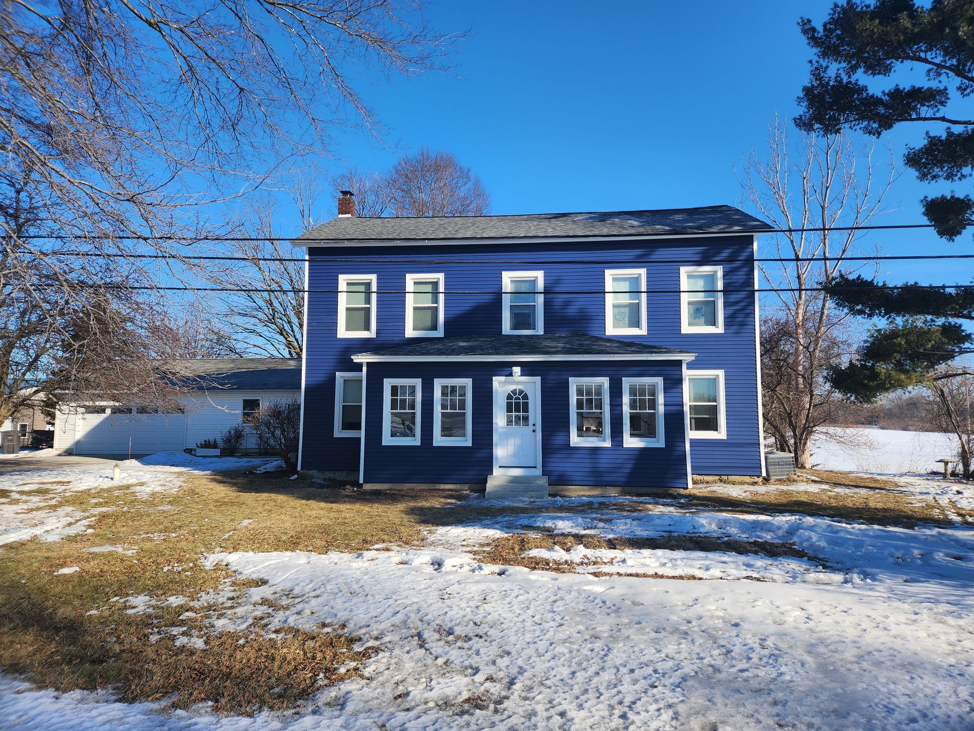 Two-story blue house with white trim against a clear, sunny sky, fronted by a snow-covered yard.