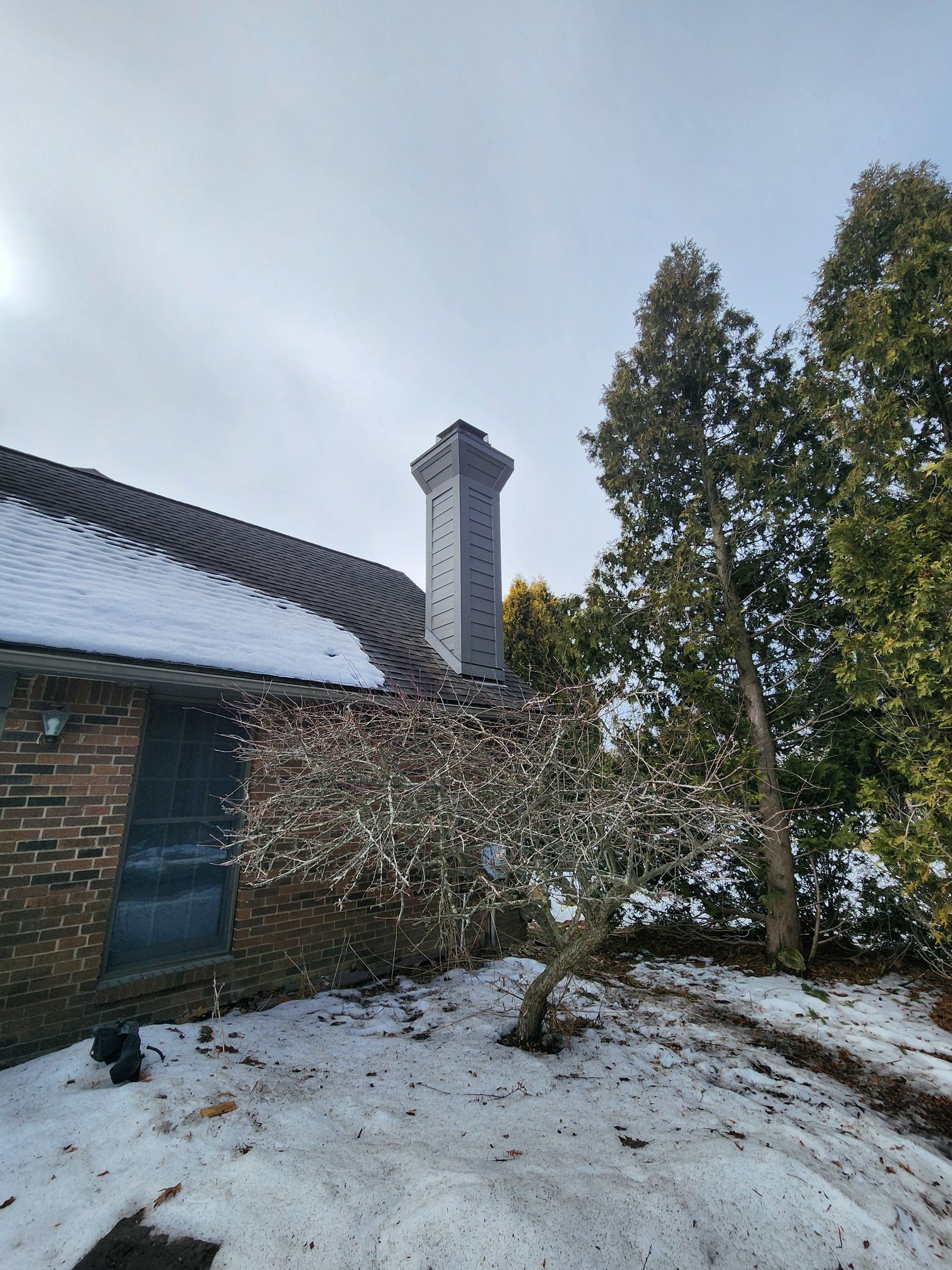 Snowy rooftop with chimney, brick building, and evergreen trees under a cloudy sky.