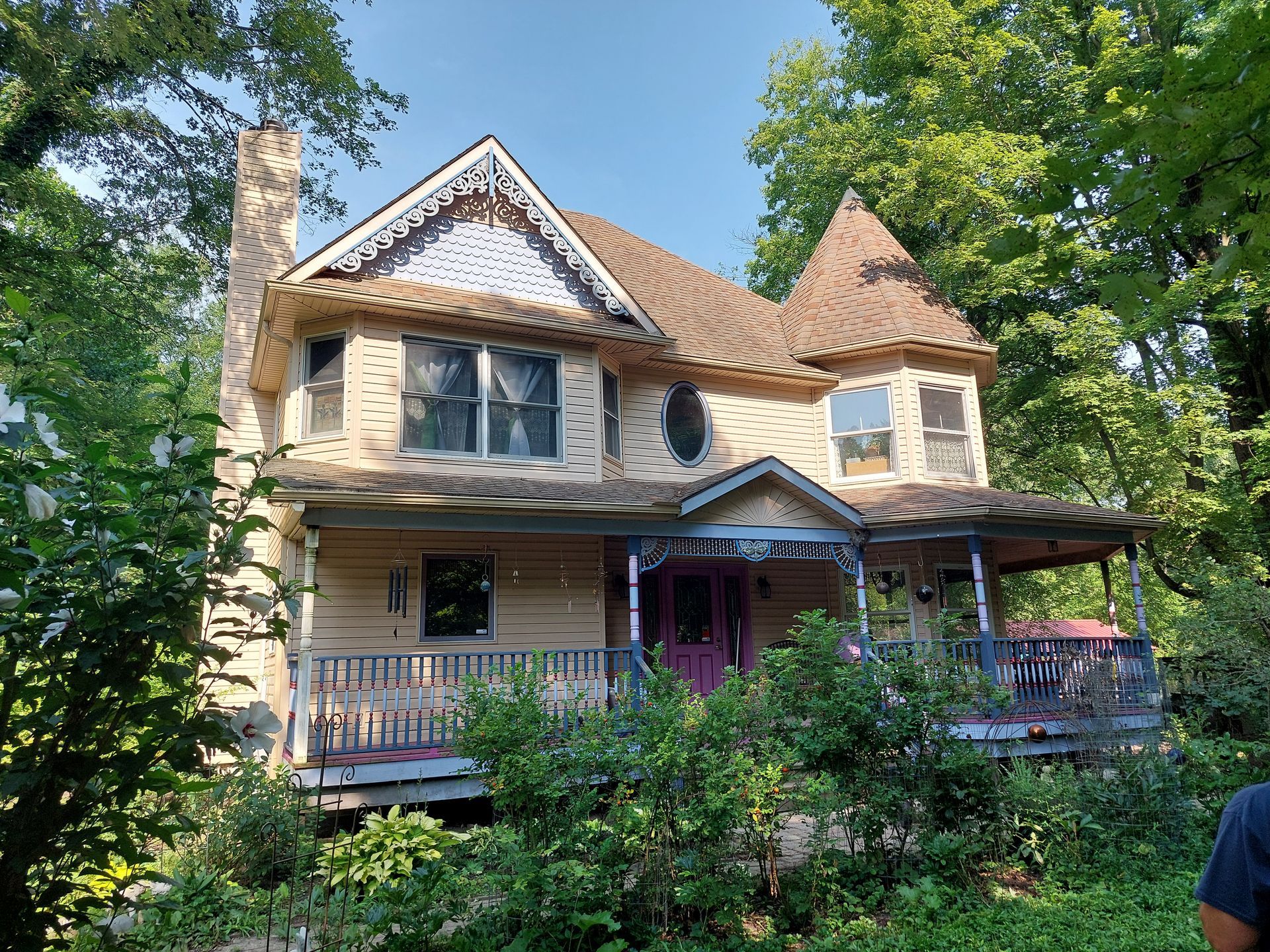 Victorian-style house with tan siding, turret, porch, and a purple door, surrounded by lush green trees.