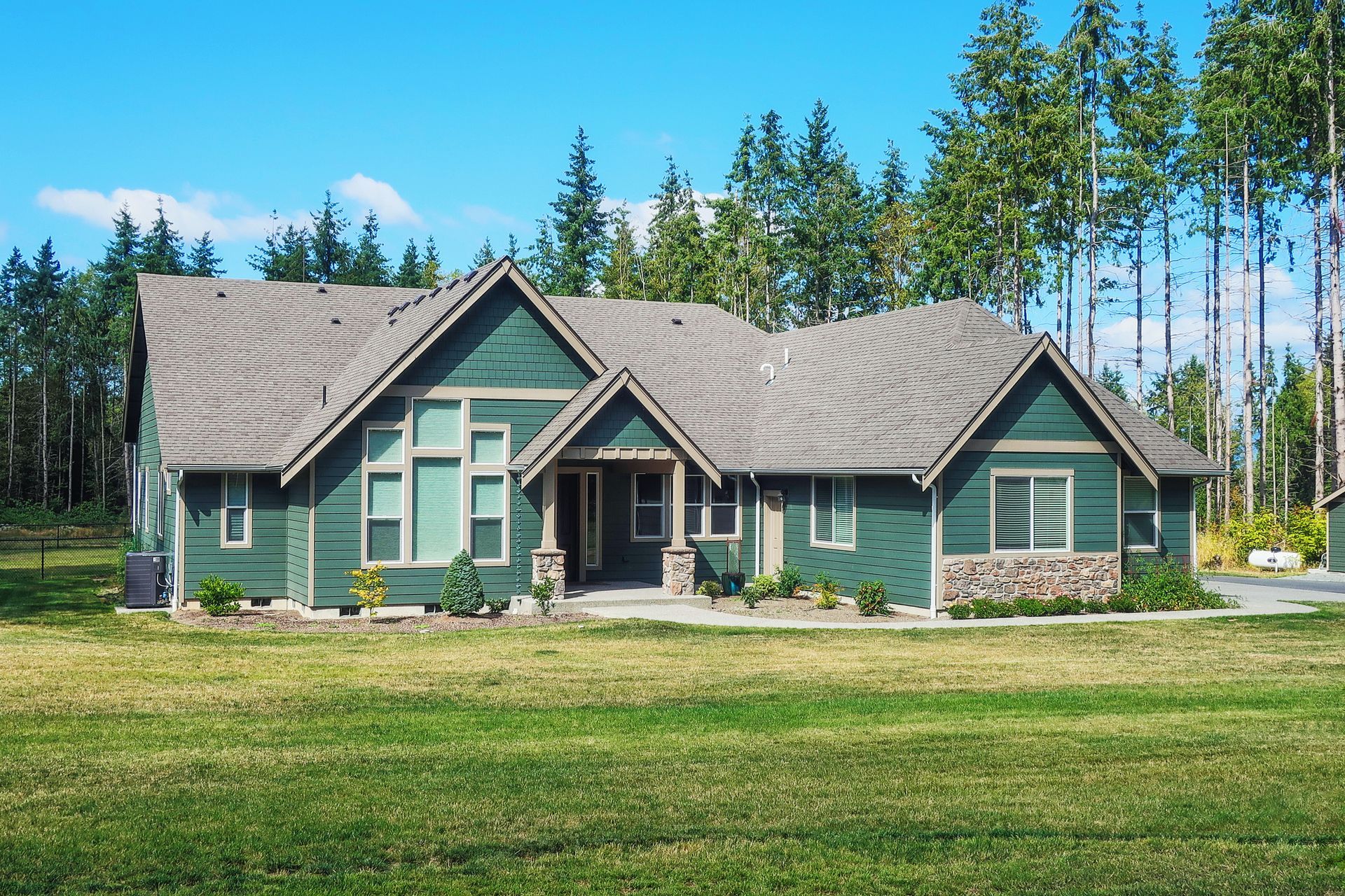 Green house with gray roof and stone accents, set against a backdrop of trees and a blue sky.