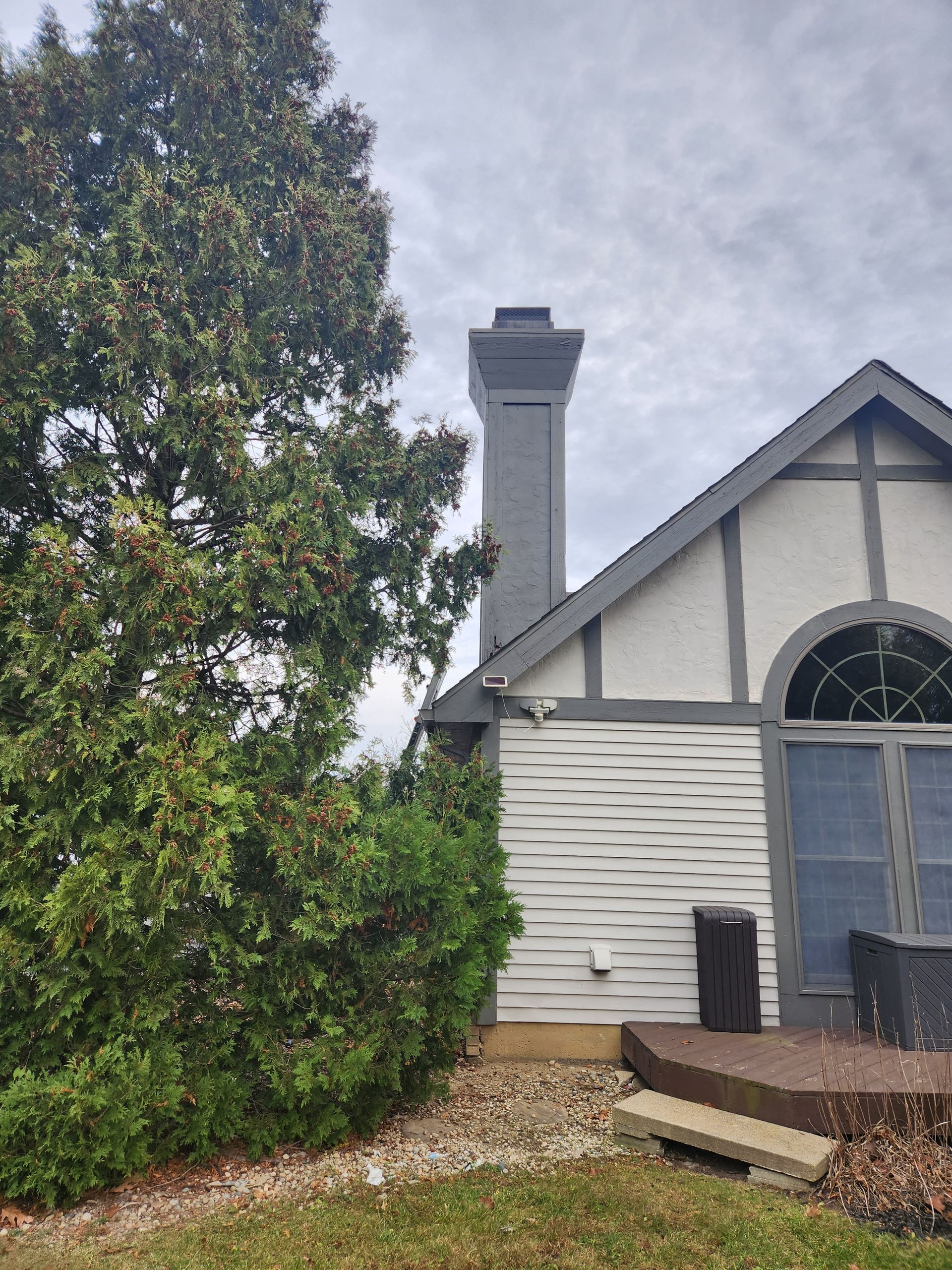 Gray chimney on a house with a white exterior and a large evergreen tree. Overcast sky.