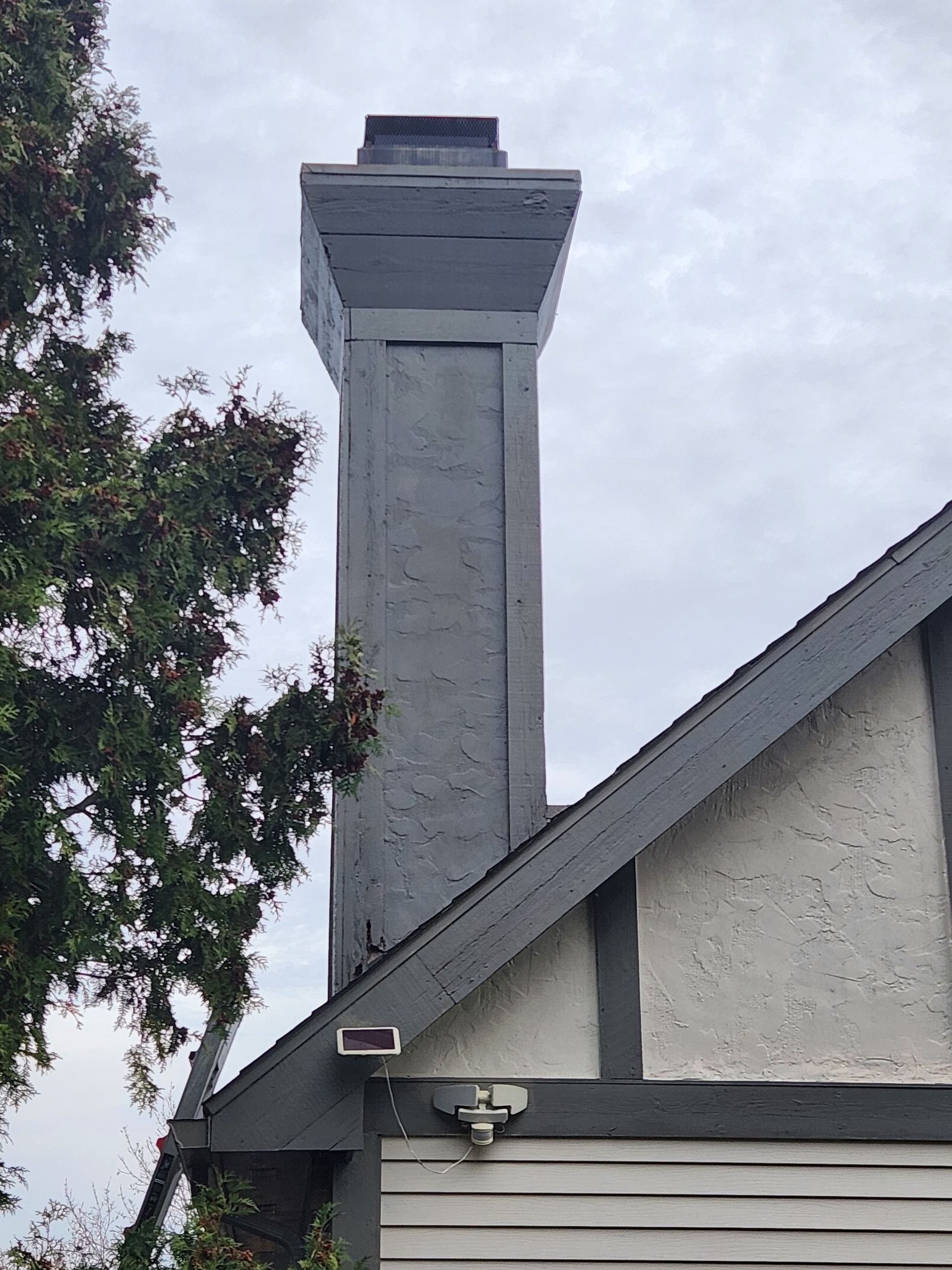Gray chimney atop a house with gray roof and white siding; cloudy sky backdrop.