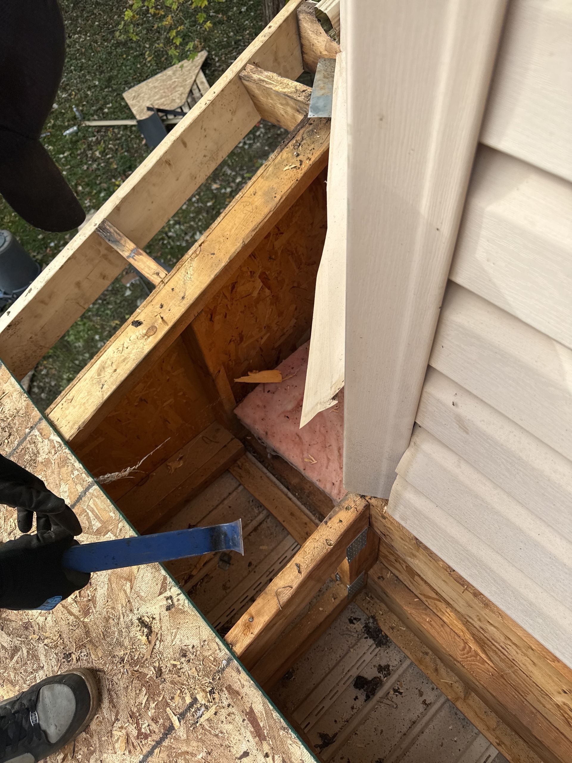 Construction worker using a pry bar to remove wood from a roof corner. Brown wood and beige siding visible.