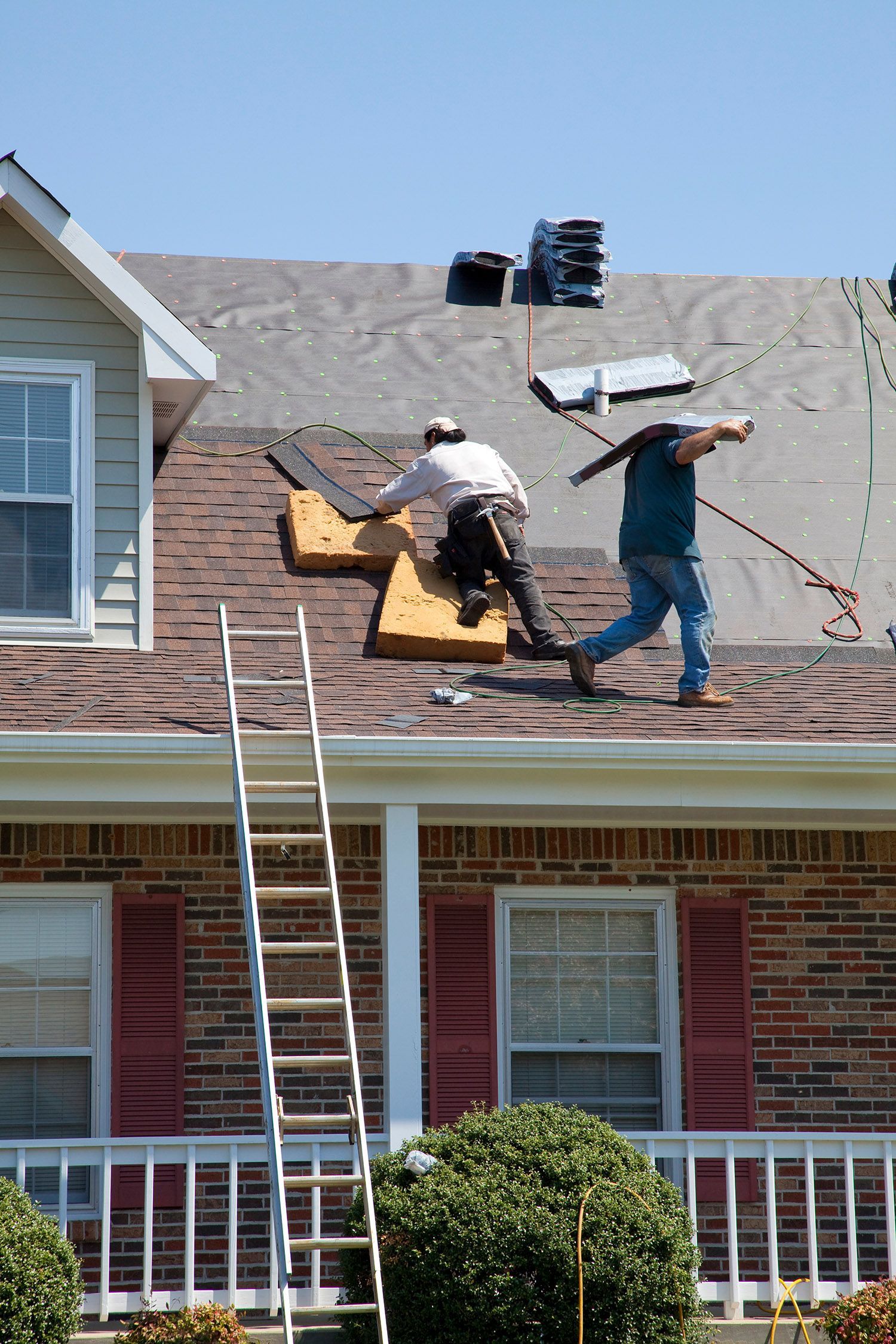 A pair of roofers install new shingles on the roof of a house, under the sun.