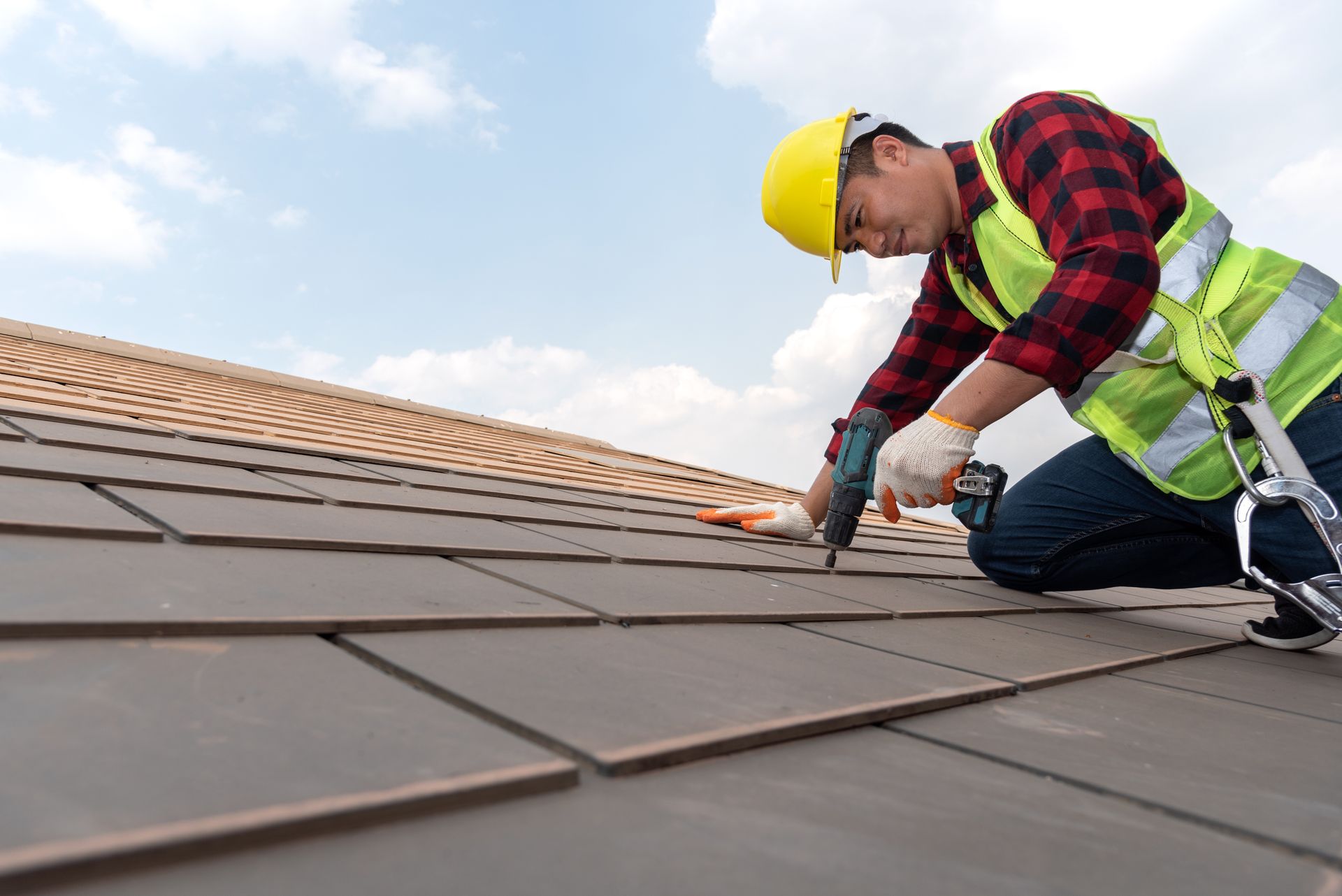 Roofer in hard hat and safety vest installing asphalt shingles on residential roof with drill.