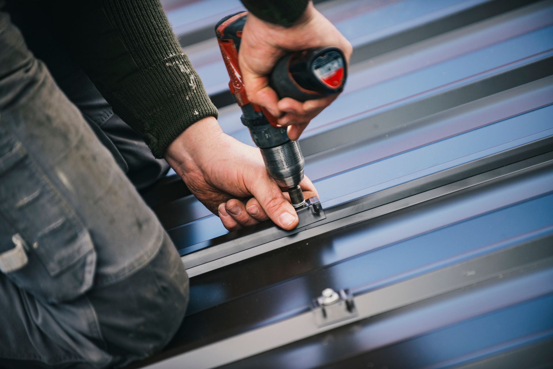 Person using a drill to fasten a metal roof panel. They are kneeling, and the roof panels are blue-gray.