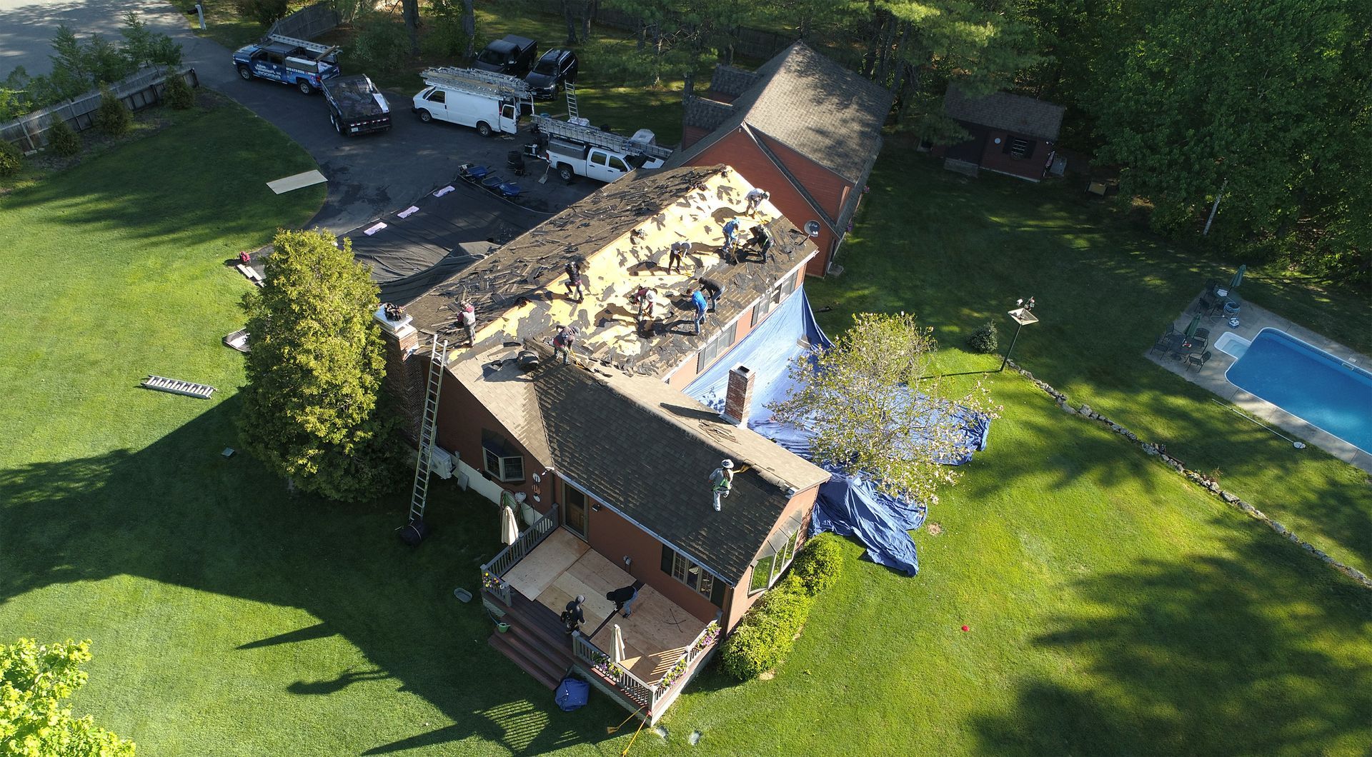 Aerial view of a house undergoing roof replacement with workers, surrounded by green grass and a swimming pool.