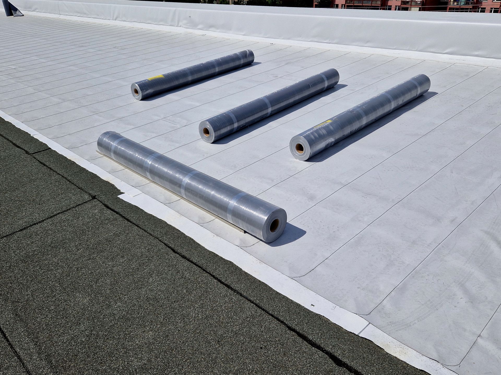 Five rolls of roofing material on a white membrane-covered roof. The edge reveals a dark gray, textured surface.