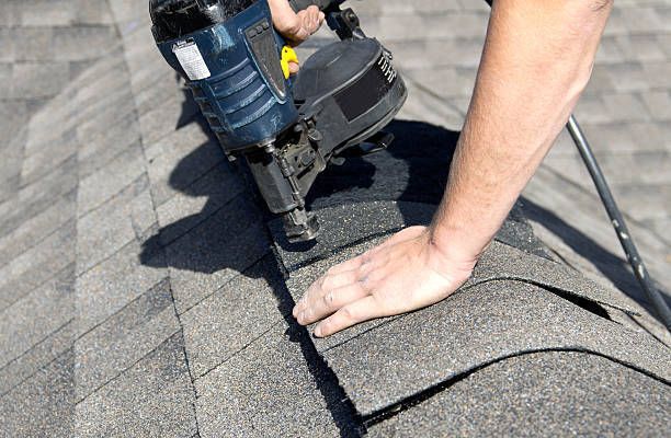 Person nailing shingles to a gray roof, using a pneumatic nail gun. Close-up view of hands and tool.