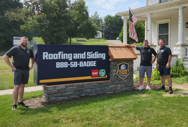Two men in front of a house with a banner advertising roofing and siding services. One is leaning on a ledge, and the other is standing next to him.