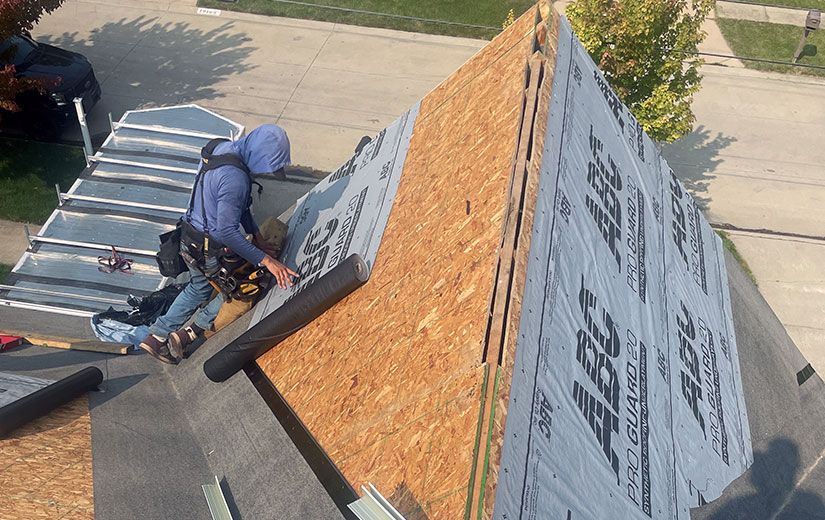 Roofer installing black underlayment on a wooden roof; he wears a safety harness.