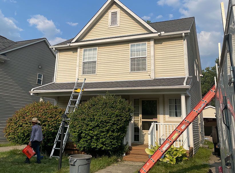 Two-story house with tan siding, a new black roof, and two ladders set up. A worker carries a red bucket.