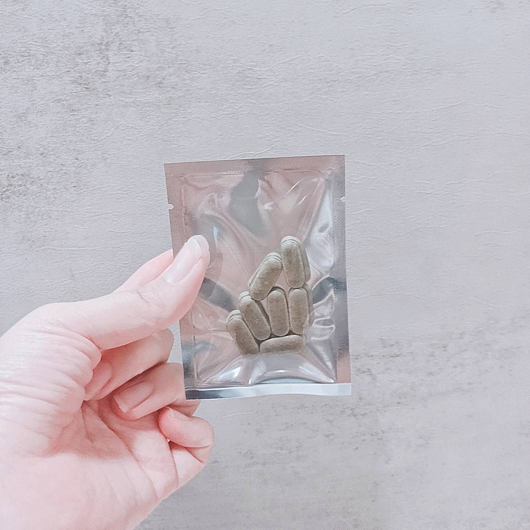 一隻手拿著一個銀色的小密封箔袋，裡面裝著幾片棕褐色的長方形藥片，背景是灰色。